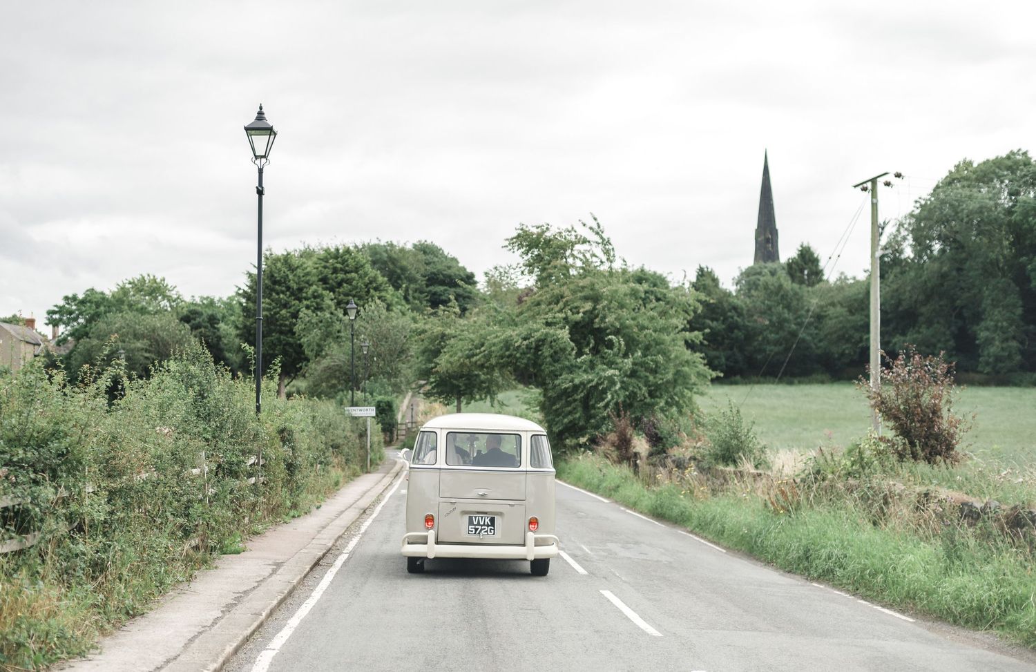 A classic white Volkswagen van driving down a country road with a lamppost in view.