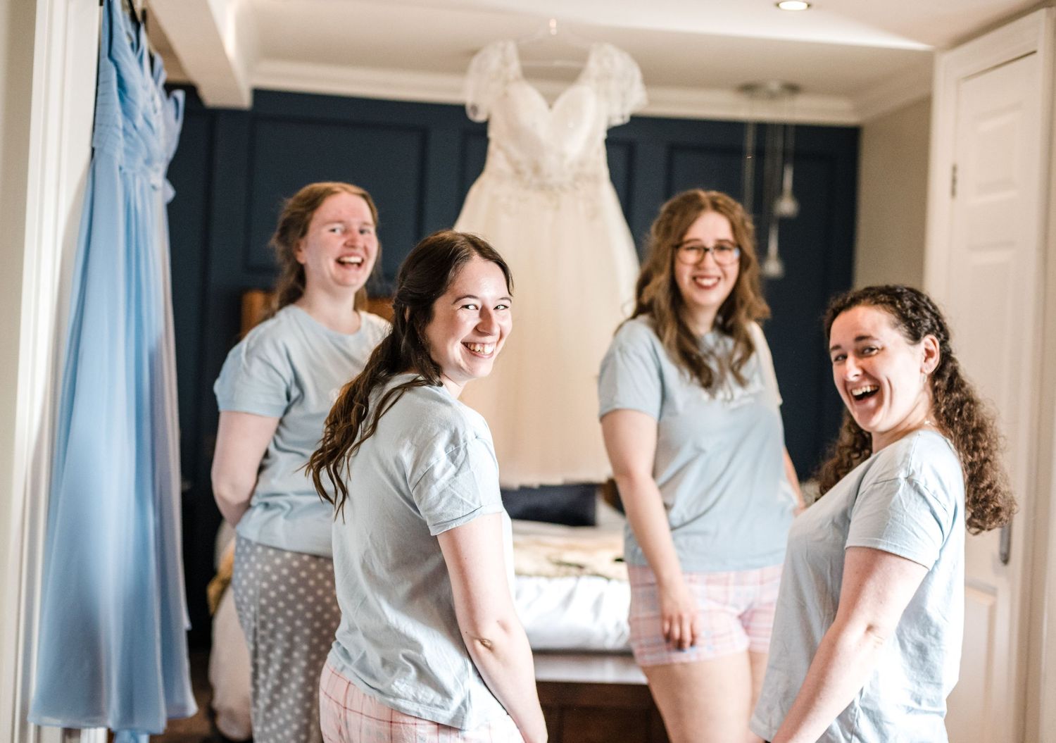 A group of bridesmaids in matching outfits share a joyful moment in a dressing room with a wedding gown displayed.