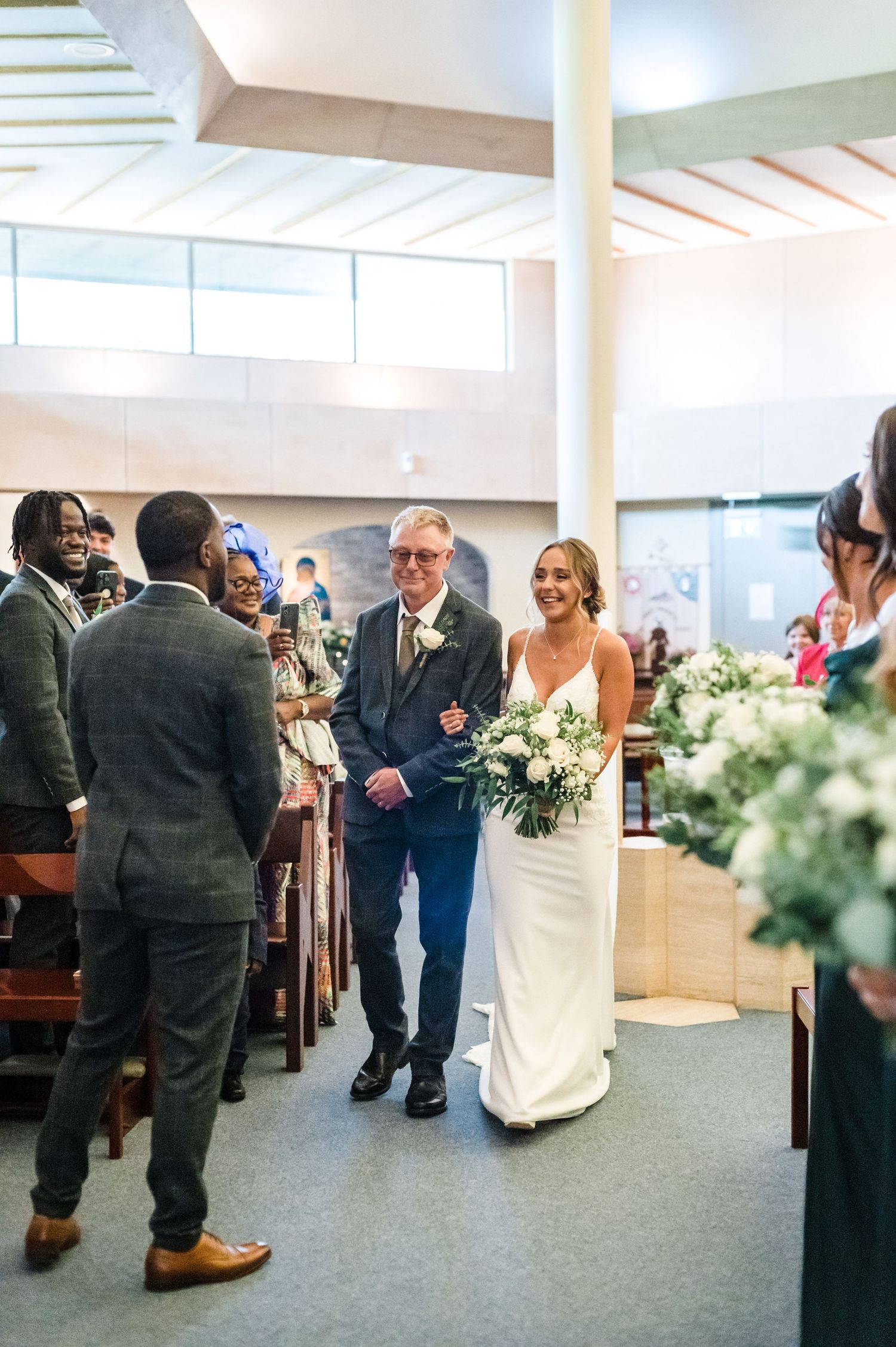 A joyful wedding procession walks down the aisle of a modern church with white walls and high windows.