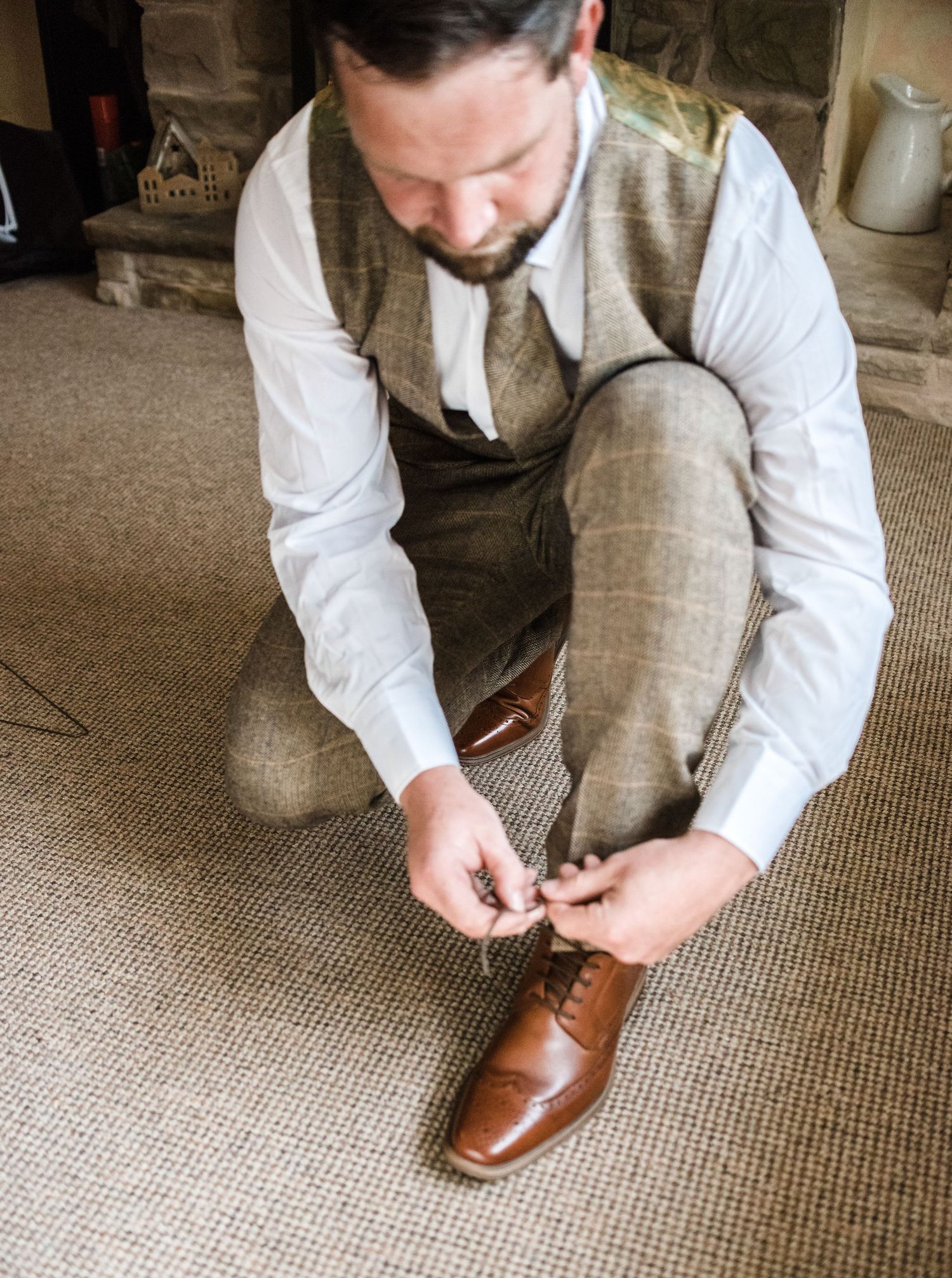 A person wearing a brown tweed vest and white shirt tying brown leather dress shoes on carpet.