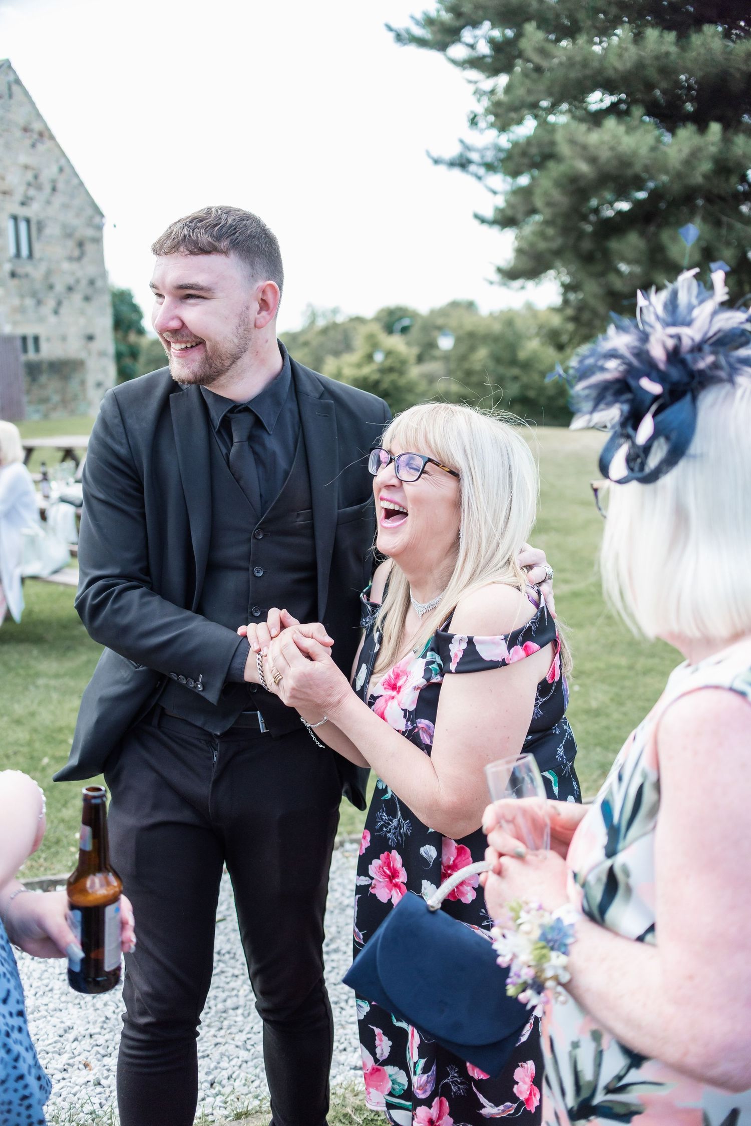 Wedding guests share a candid laughing moment while holding drinks at an outdoor reception.