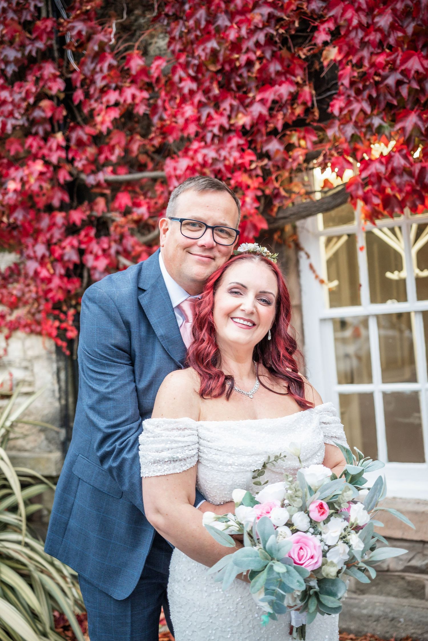 A wedding couple poses against a wall covered in vibrant red autumn foliage while holding a pink and eucalyptus bouquet.