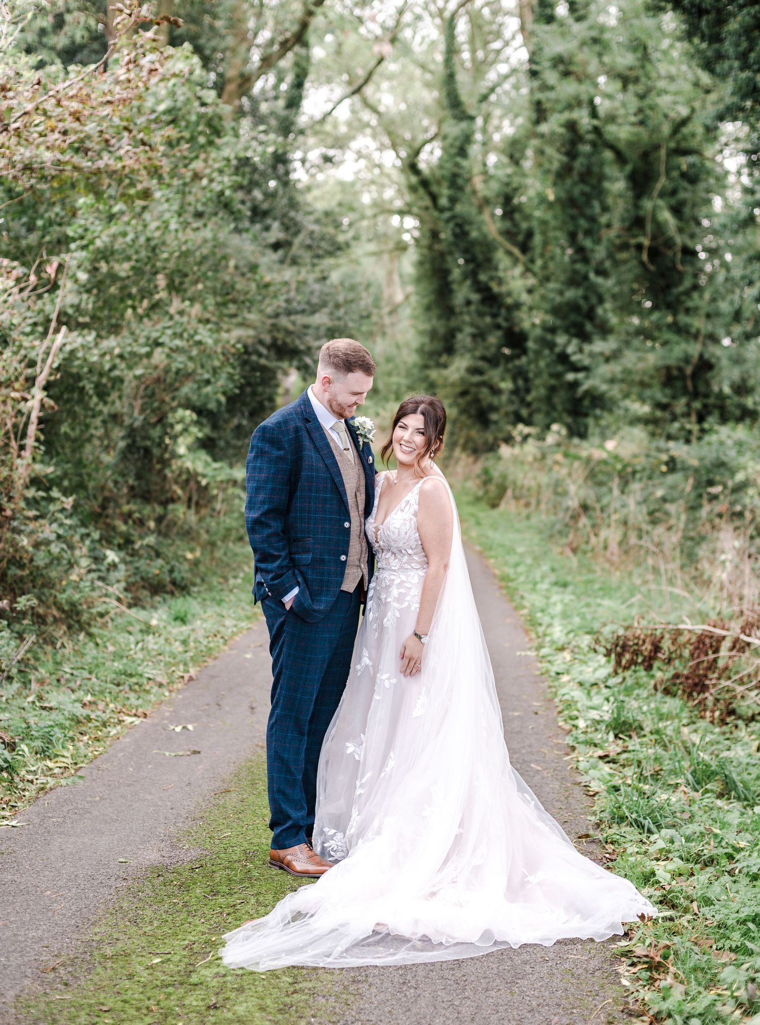 Couple stands on garden path showcasing the bride's flowing lace train and groom's navy suit.