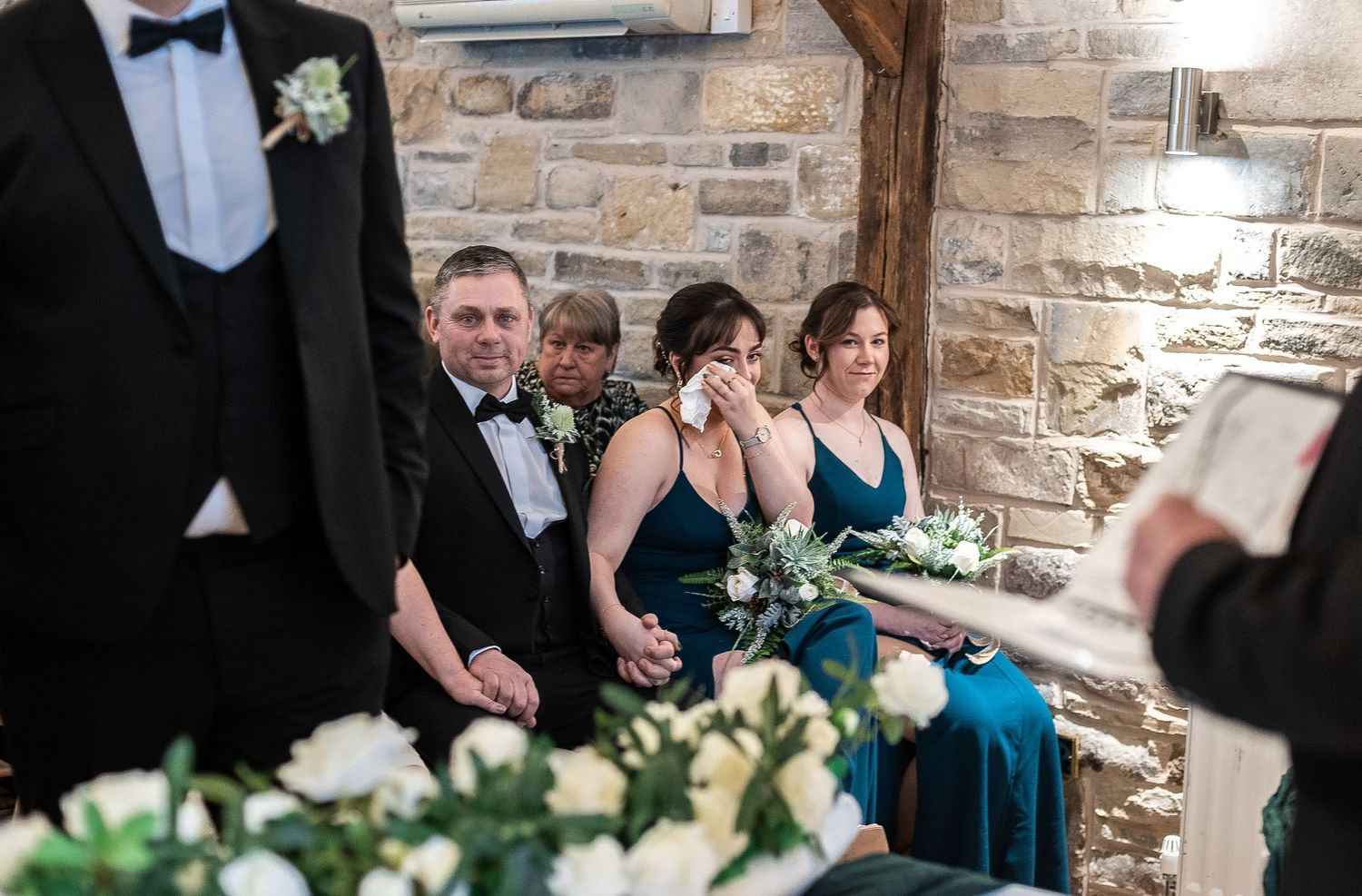 Wedding ceremony attendees seated in a rustic stone venue with elegant white floral arrangements.