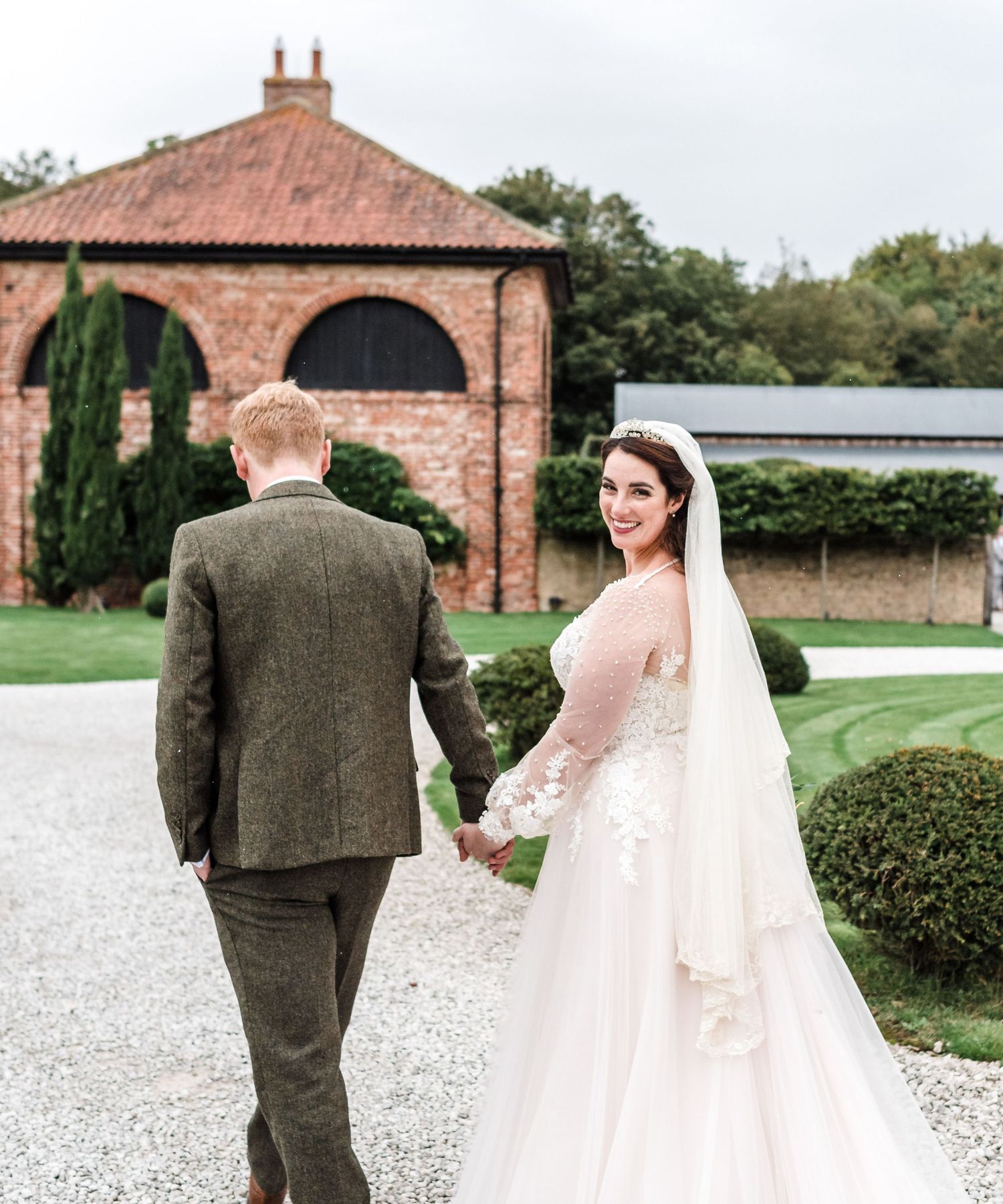 Wedding couple poses in white gown and tweed suit near historic brick building with arched windows.