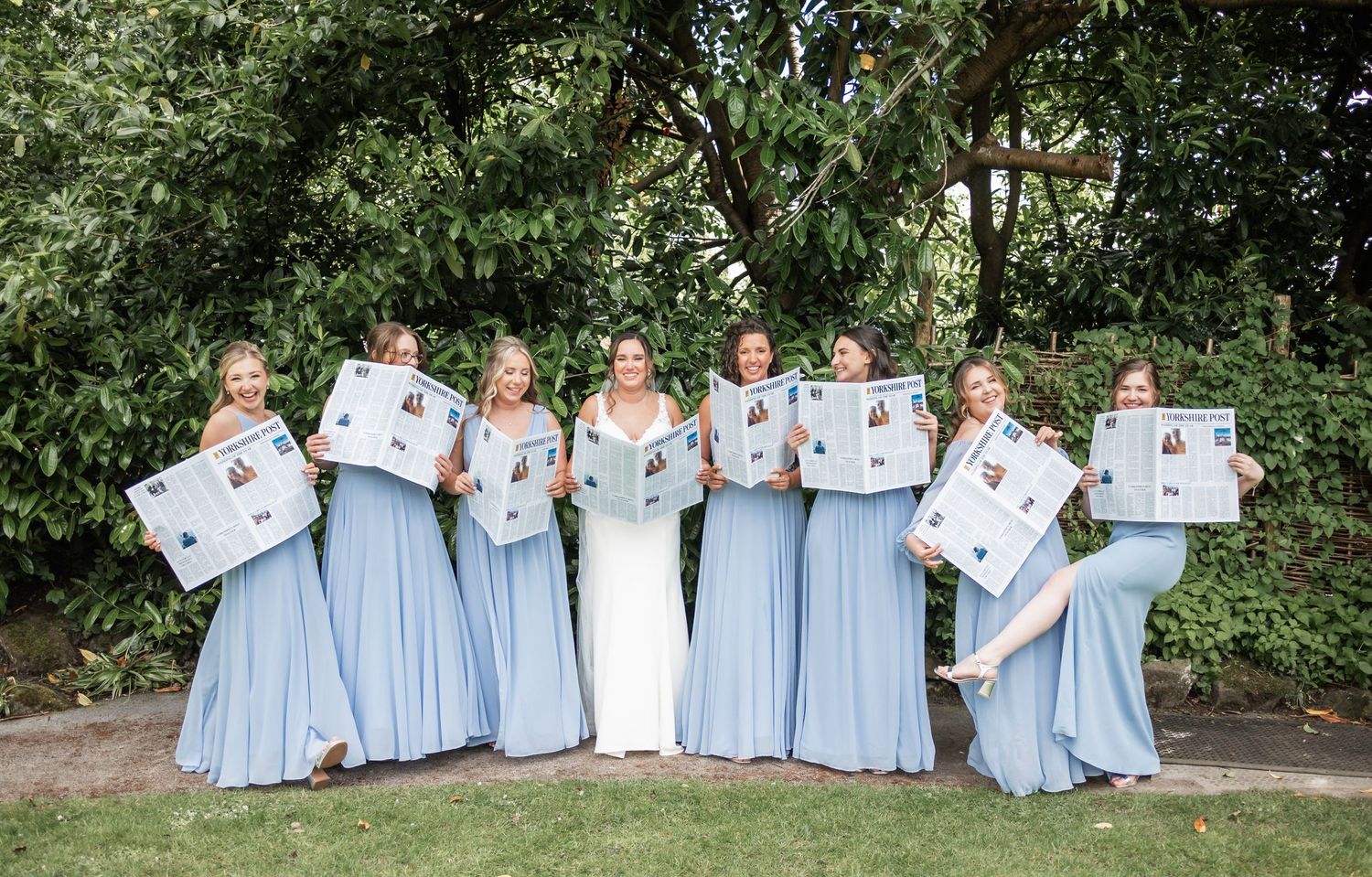 A bridal party in light blue dresses holding up white photo frames in a garden setting.