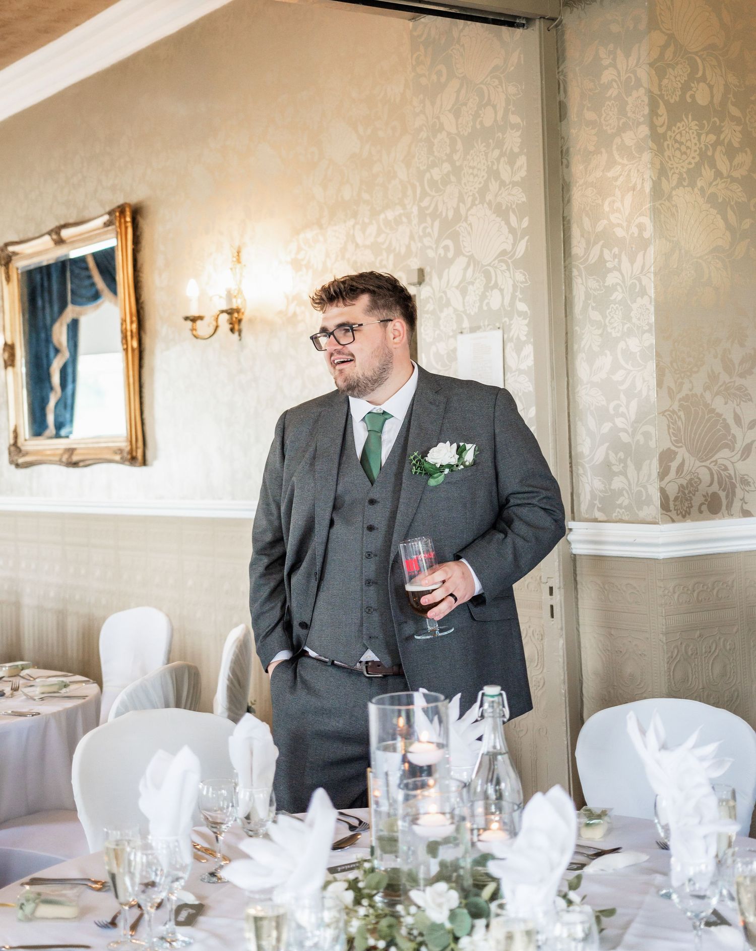 Wedding reception setup with gray-suited figure holding drink beside white-covered tables.