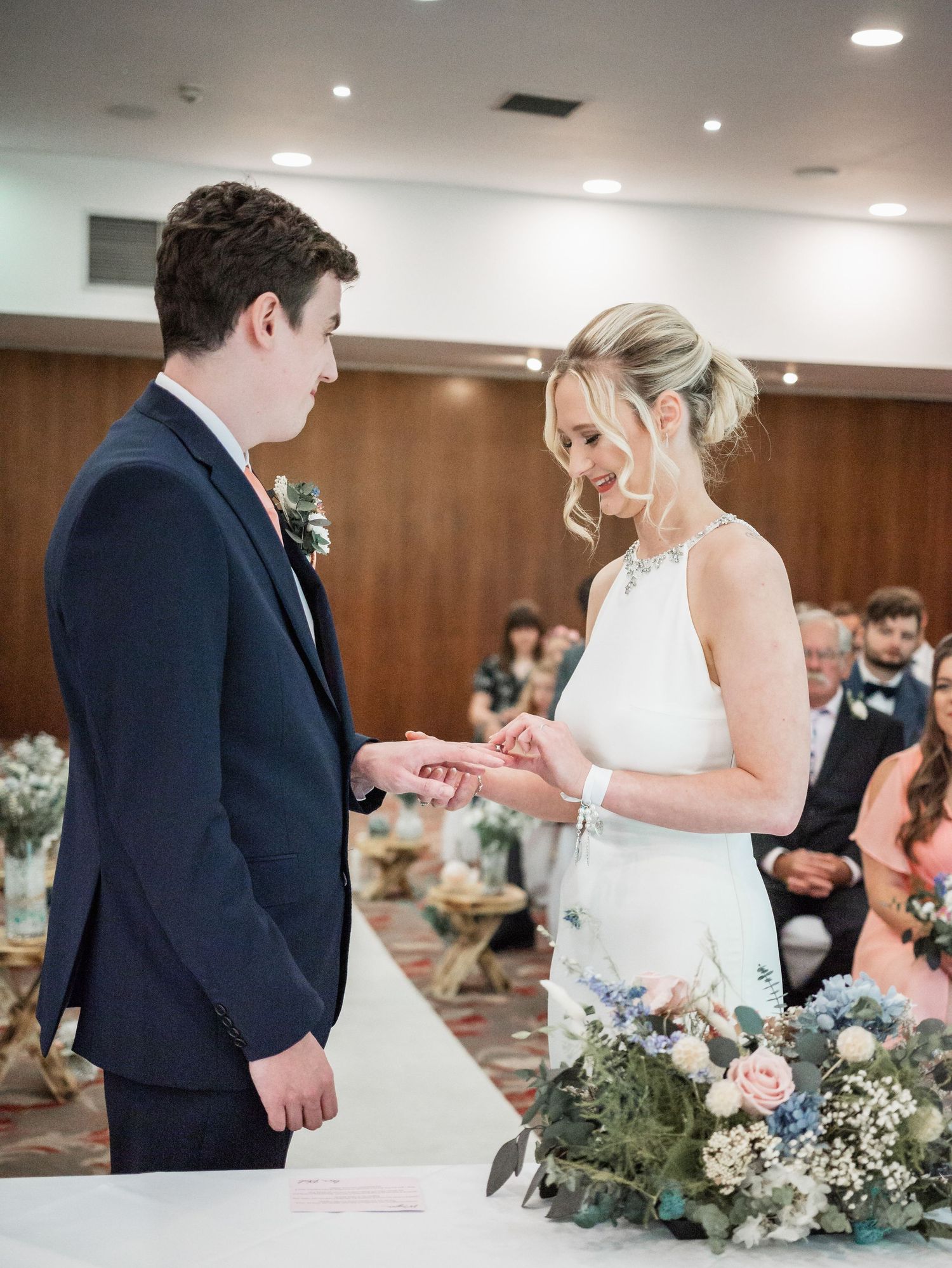 A couple exchanges rings during their wedding ceremony surrounded by floral arrangements and guests.
