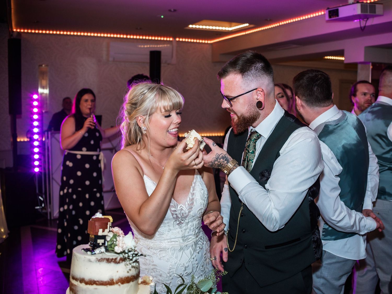 Wedding cake cutting moment captured under romantic string lights and pink ambient lighting.
