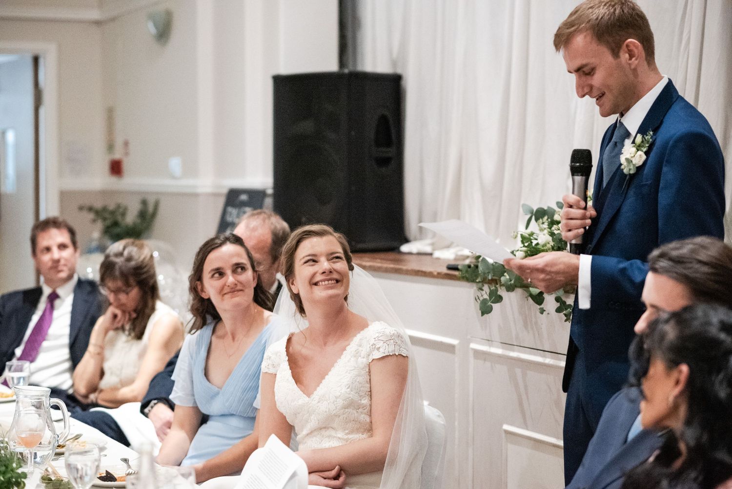 Wedding reception guests seated at long tables in an elegant white room with natural light.