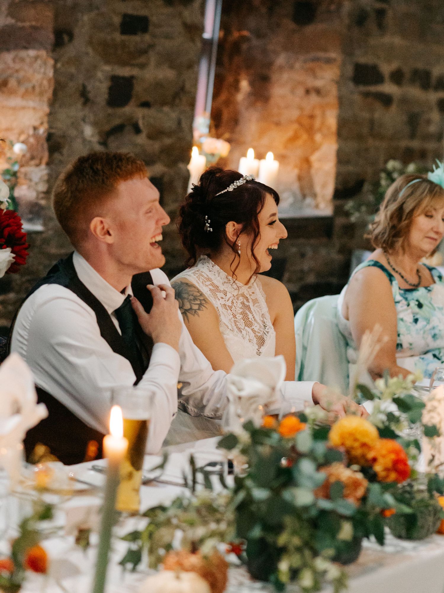 Wedding reception dinner table with orange and yellow floral centerpieces and stone wall backdrop.