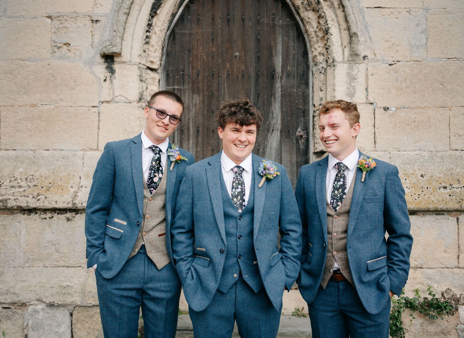 Groomsmen in matching navy blue suits pose in front of stone church archway.