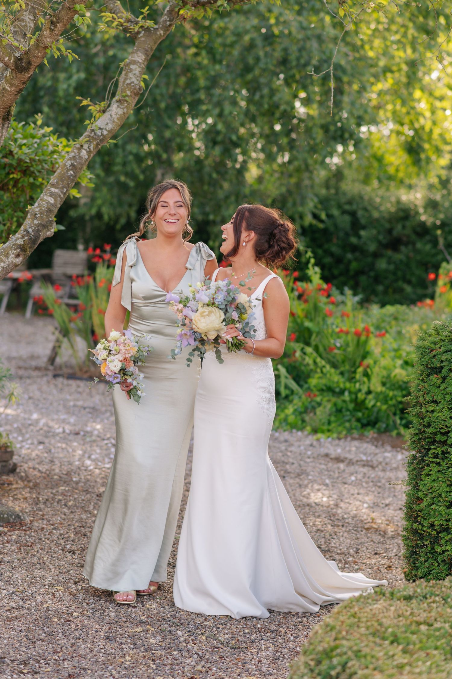 Two people in white wedding dresses share a joyful moment together in a garden setting with lush greenery.