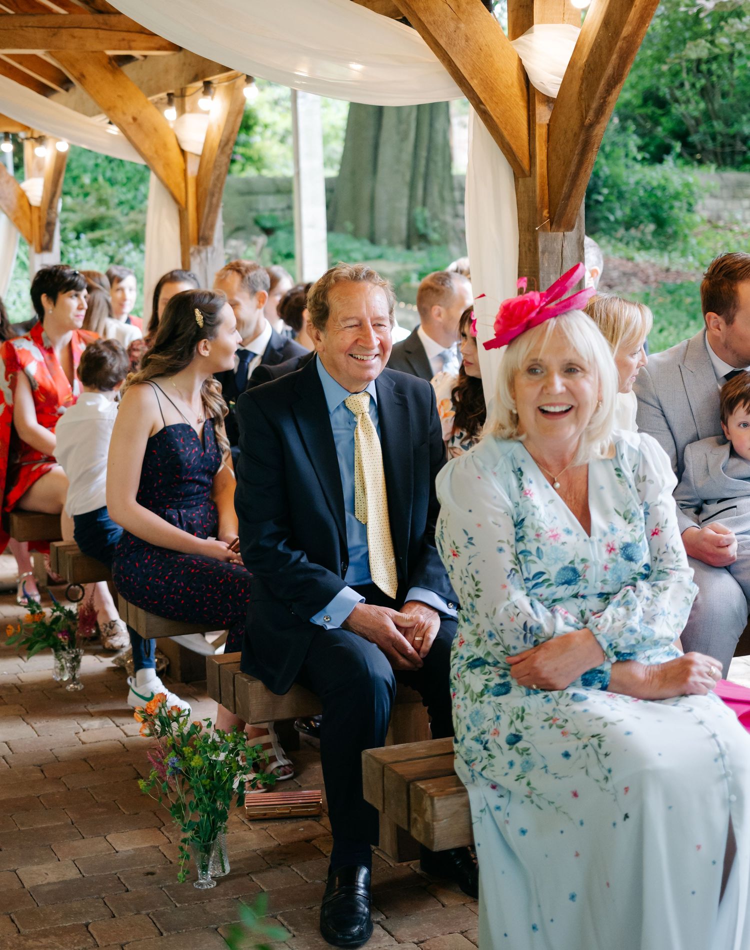 Wedding guests seated at an outdoor covered venue with wooden beams and floral centerpieces.