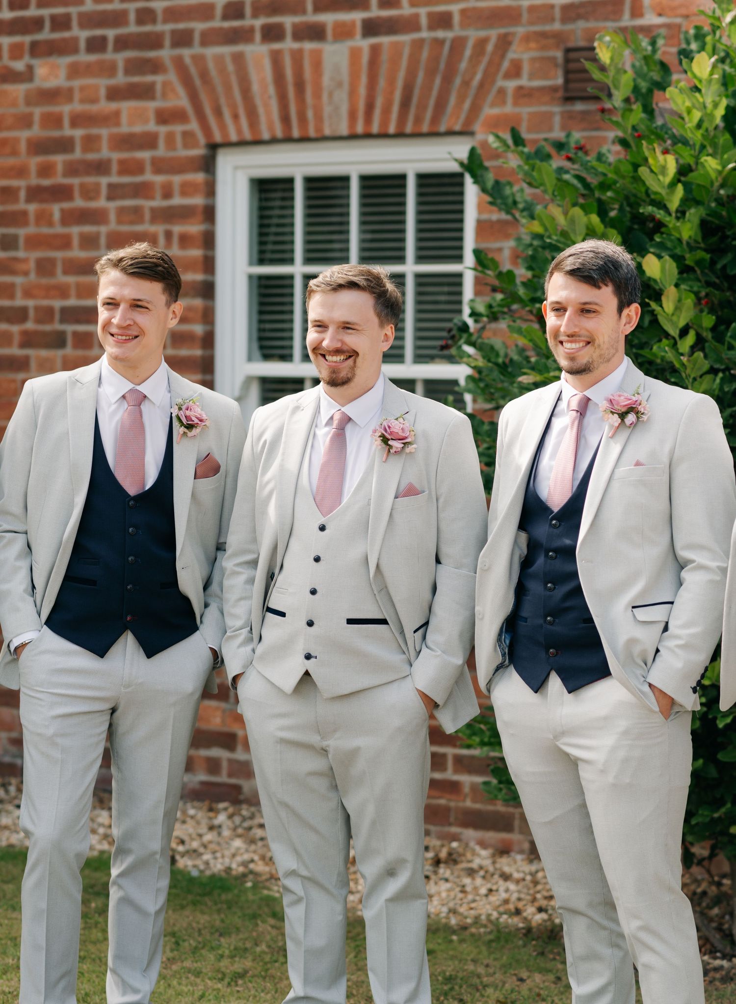 Three groomsmen in light gray suits and pink ties stand together smiling outside a brick building.
