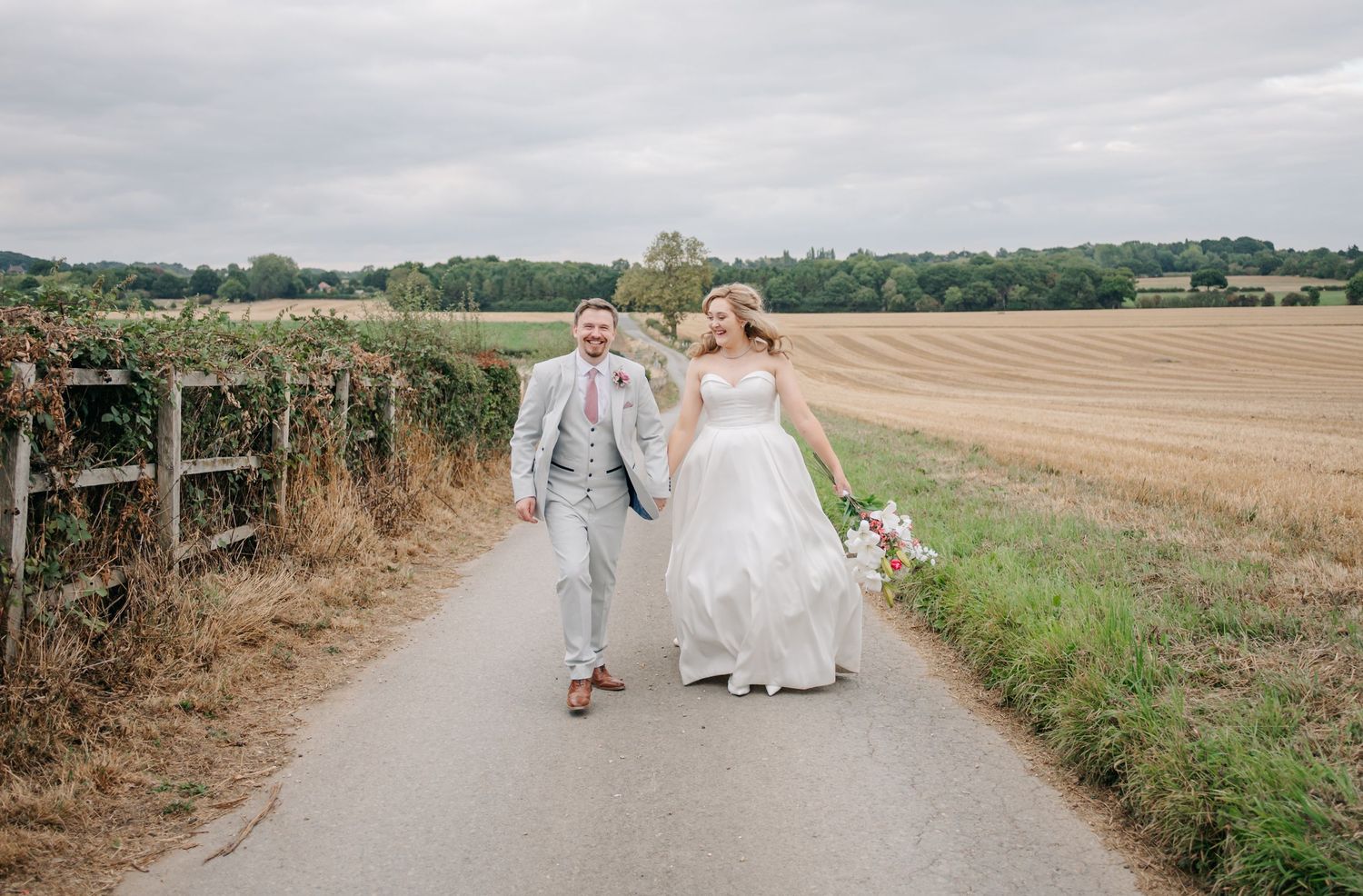 A couple walks hand in hand down a country path surrounded by summer fields.