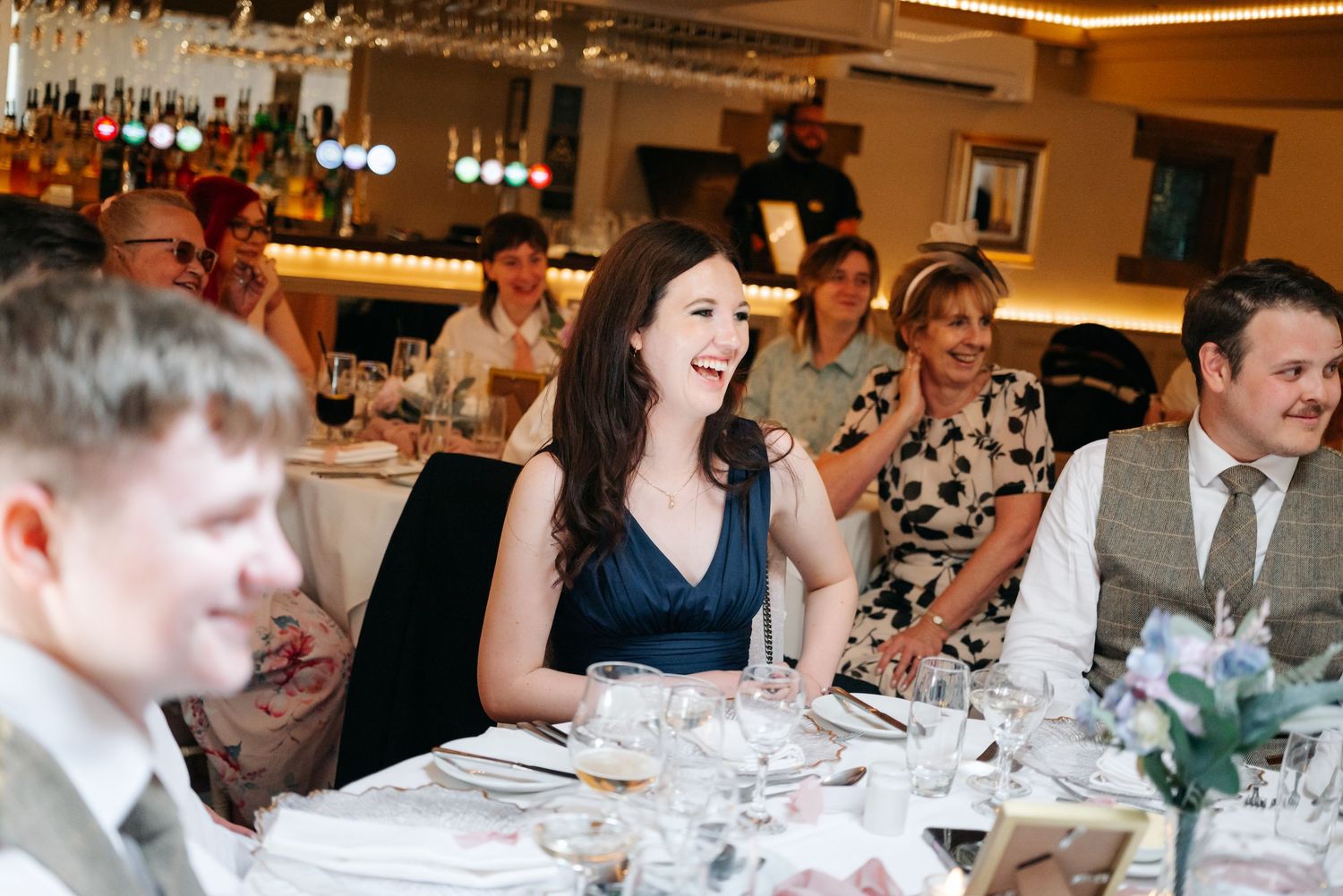 Joyful wedding guest in navy dress laughing during speeches at elegant restaurant wedding reception dinner.