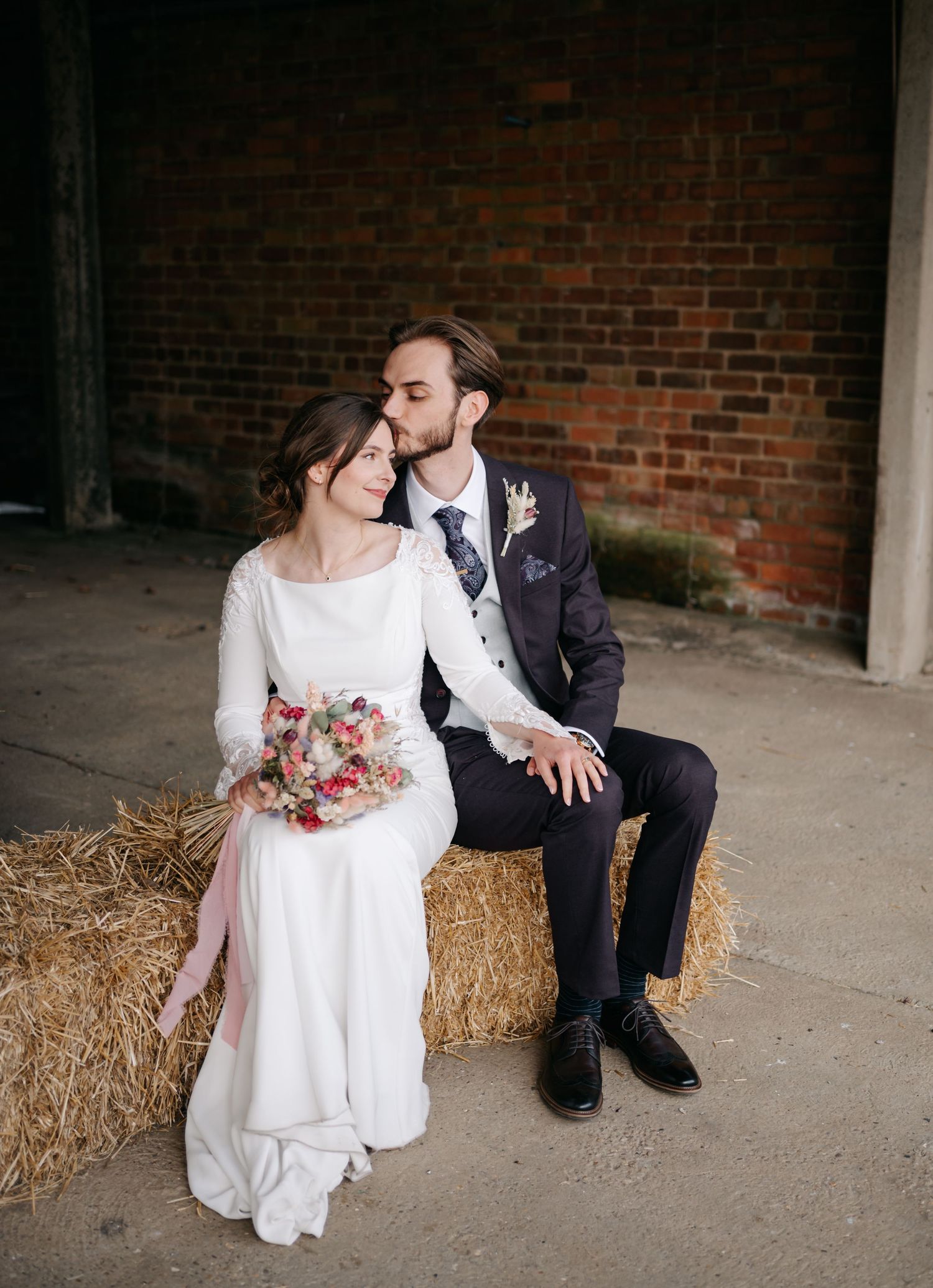 Couple sitting intimately on hay bales against a rustic brick wall backdrop.