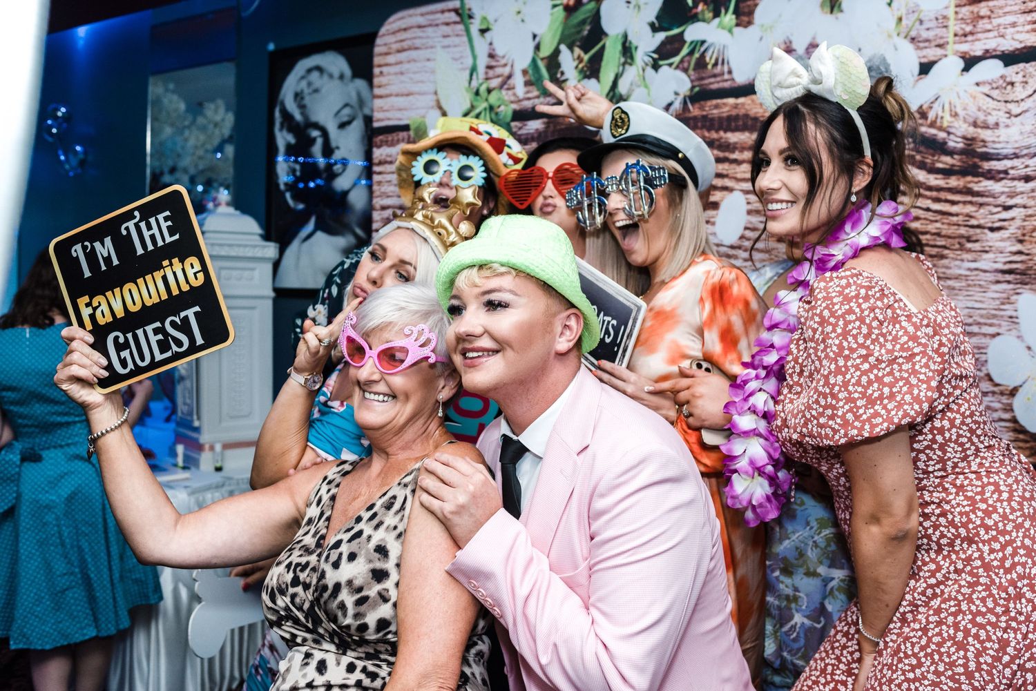 Wedding guests pose with props and colorful accessories against a floral backdrop.