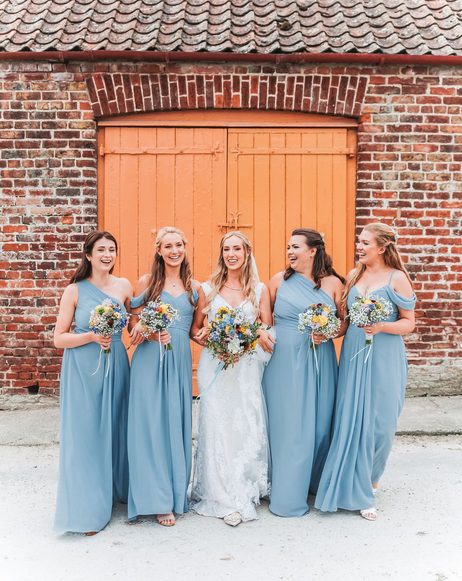 Wedding party in blue bridesmaid dresses pose against orange brick wall.