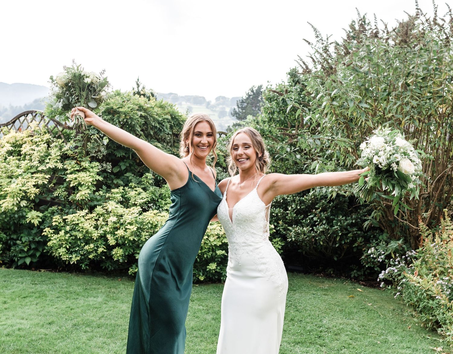 Two people in formal attire striking celebratory poses in a garden setting.