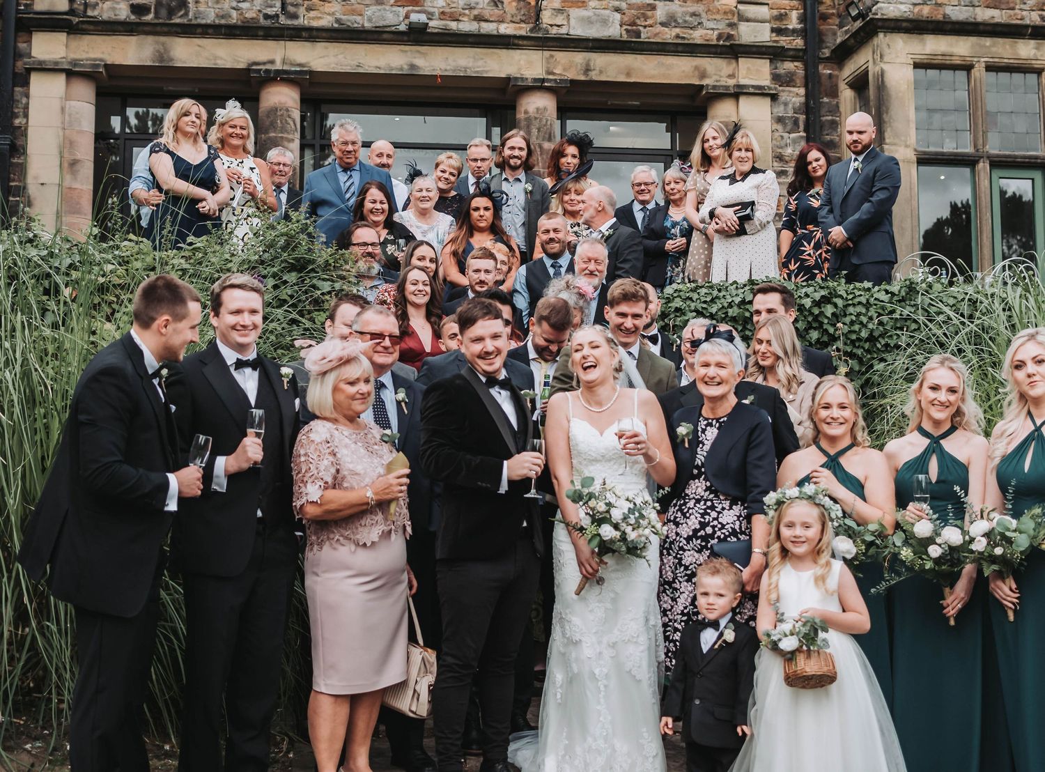 Large wedding group portrait with bridal party and guests in front of historic stone building.