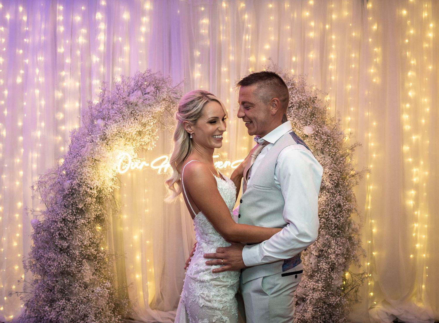 Romantic moment between newlyweds under a floral arch with fairy light backdrop.