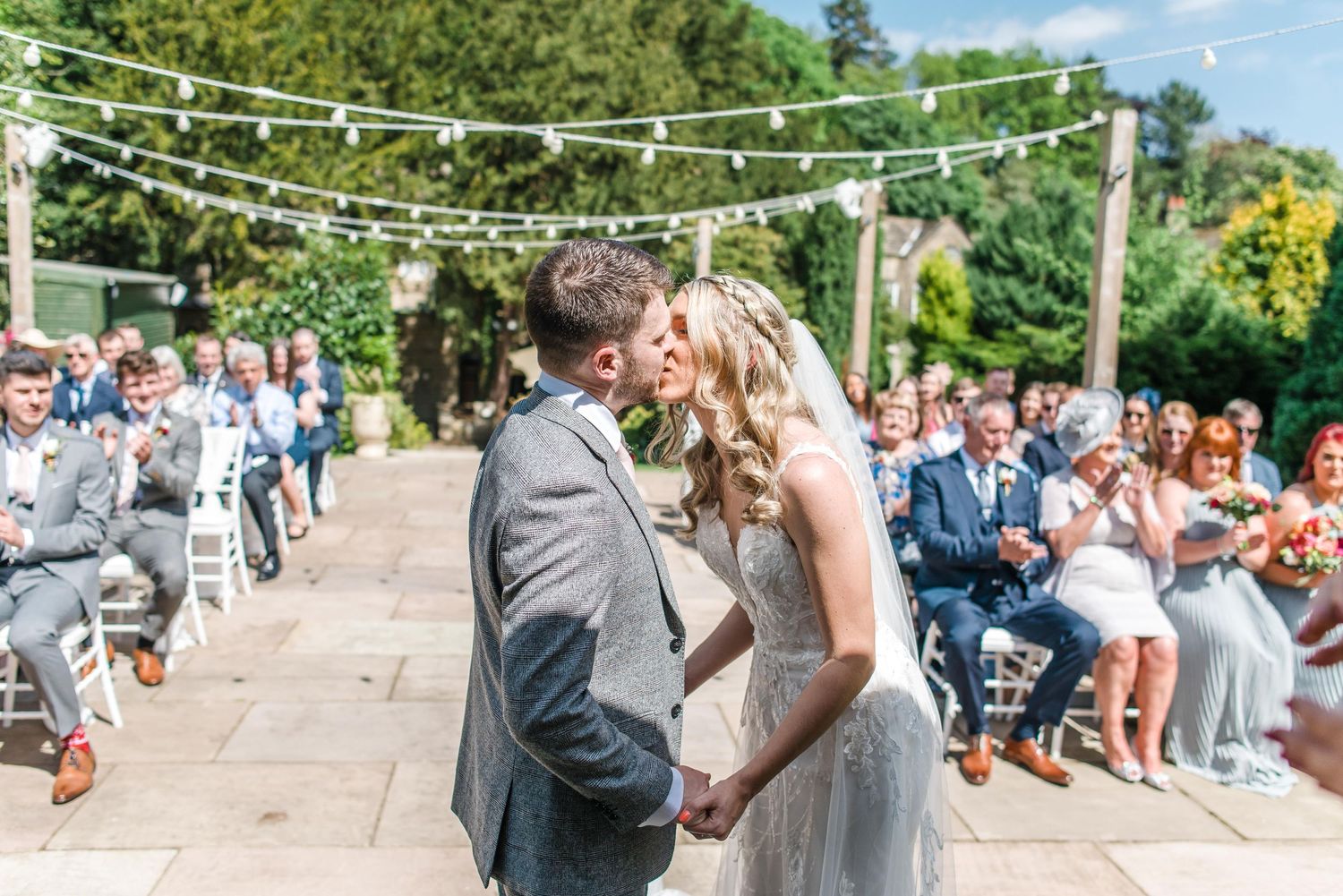 Wedding ceremony under string lights with guests seated in white chairs on stone patio.