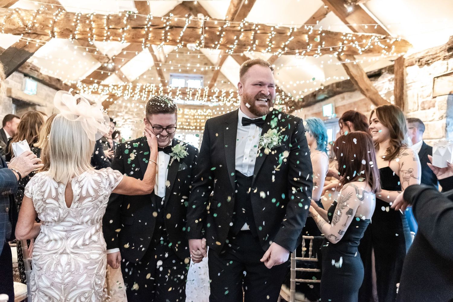 Wedding celebration with confetti falling as guests cheer in rustic venue with string lights.