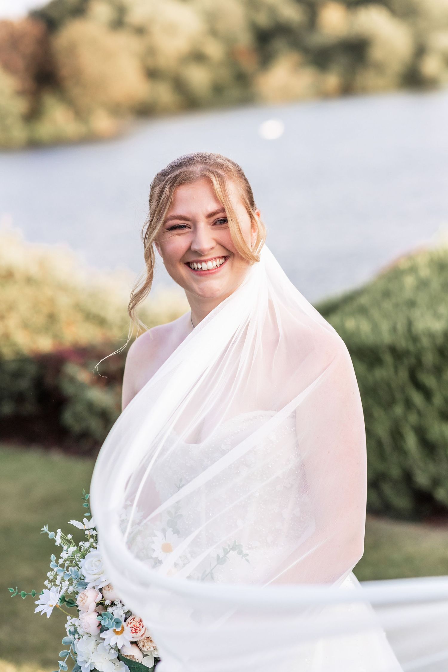 Joyful bridal portrait outdoors with flowing veil and delicate bouquet by the water.