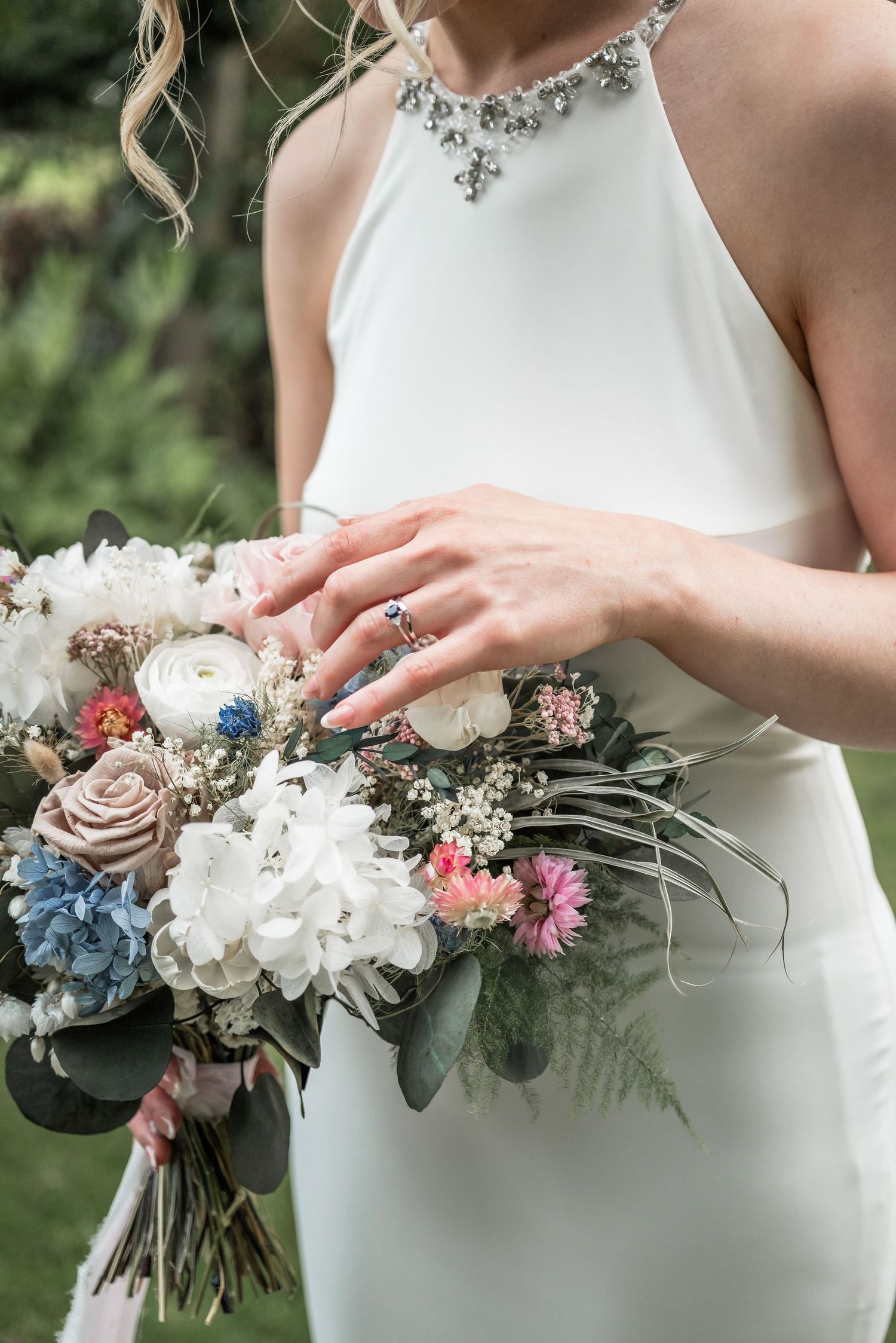 Close-up of bridal bouquet with blue and pink flowers held by hands wearing engagement ring.