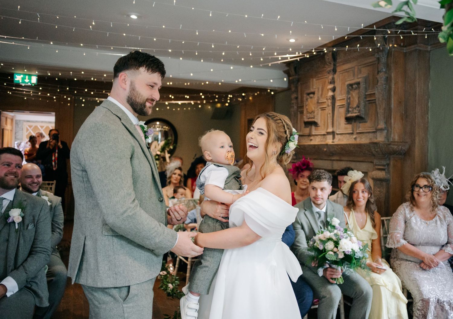 A couple sharing a tender moment with a baby during their rustic barn wedding ceremony with string lights overhead.