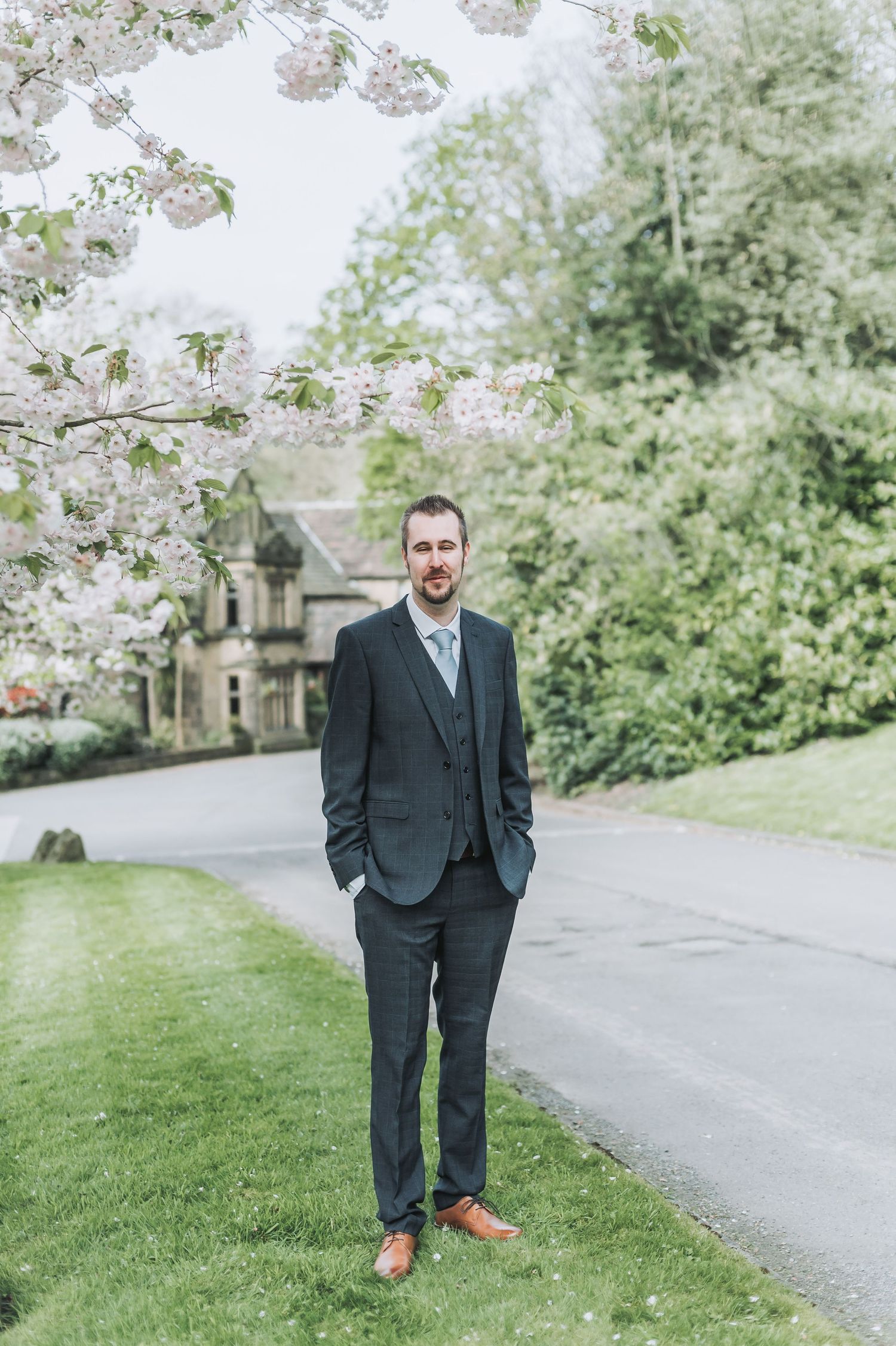 Groom in dark suit stands on lawn path with blooming cherry trees and stone building in background.