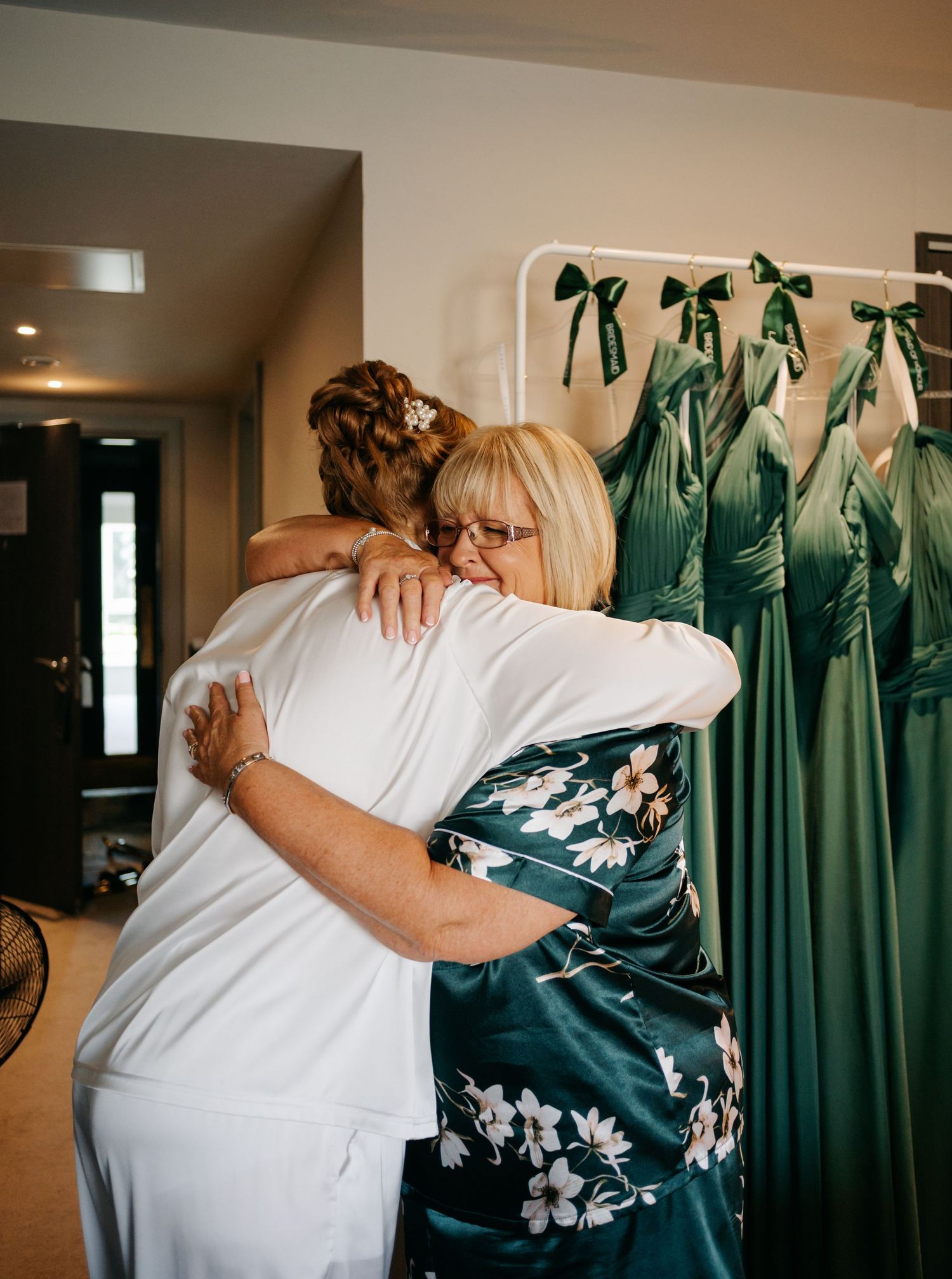 Two people share an emotional embrace while emerald green bridesmaid dresses hang in the background.