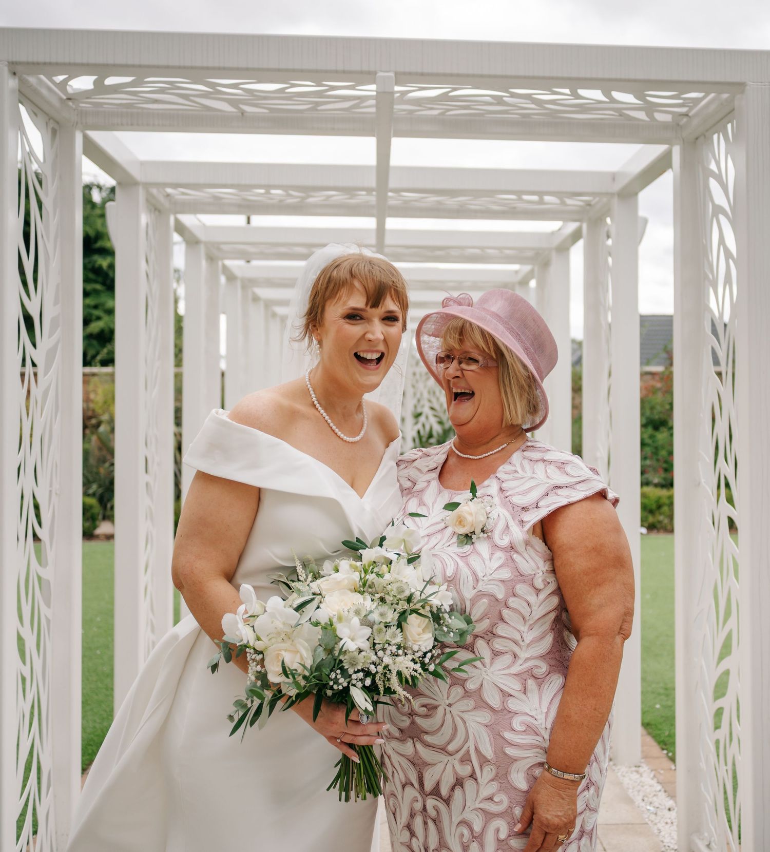 Two wedding guests share a joyful moment in a white pergola with floral bouquet.
