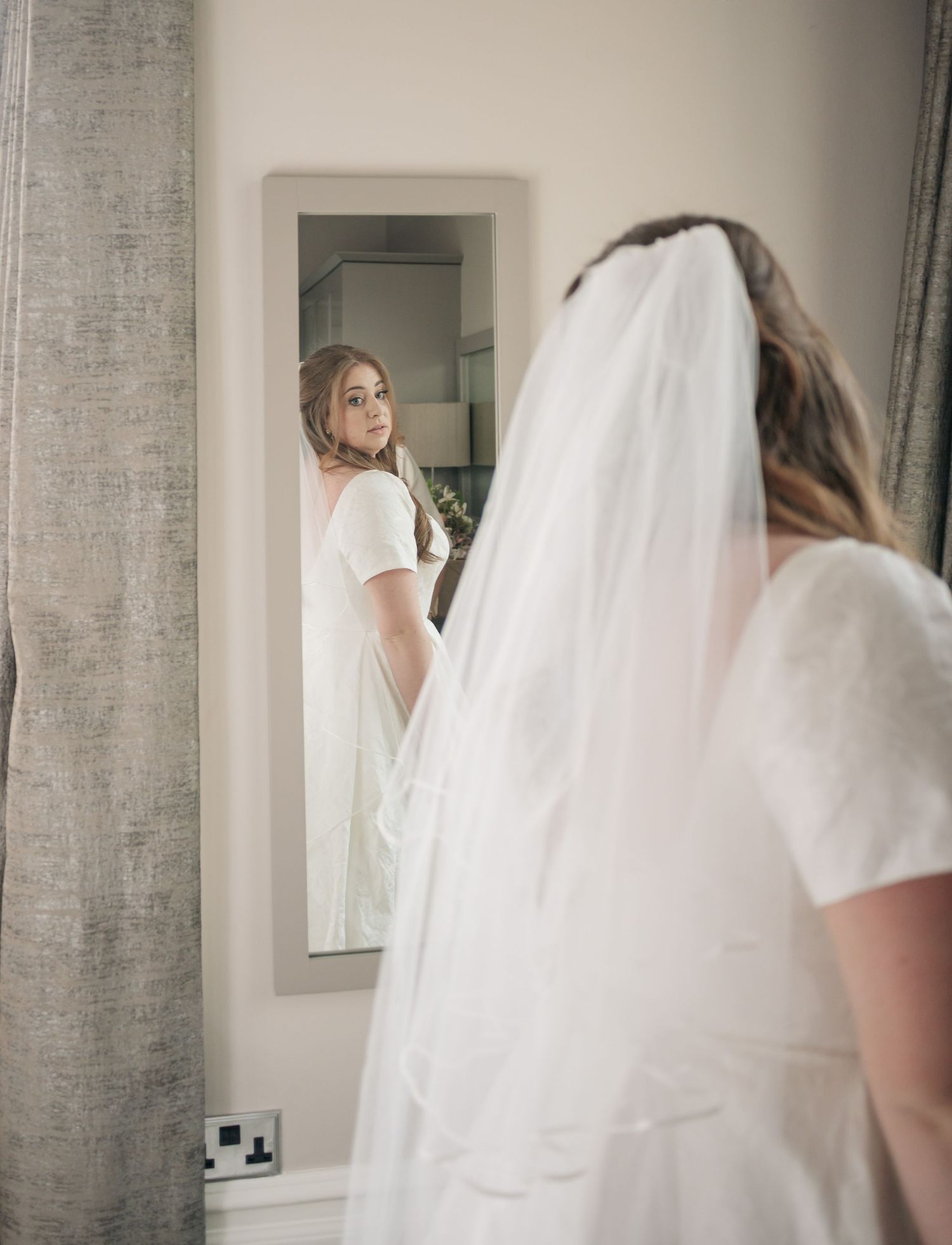 A reflection in a mirror shows a bride in a white dress and veil looking over her shoulder.