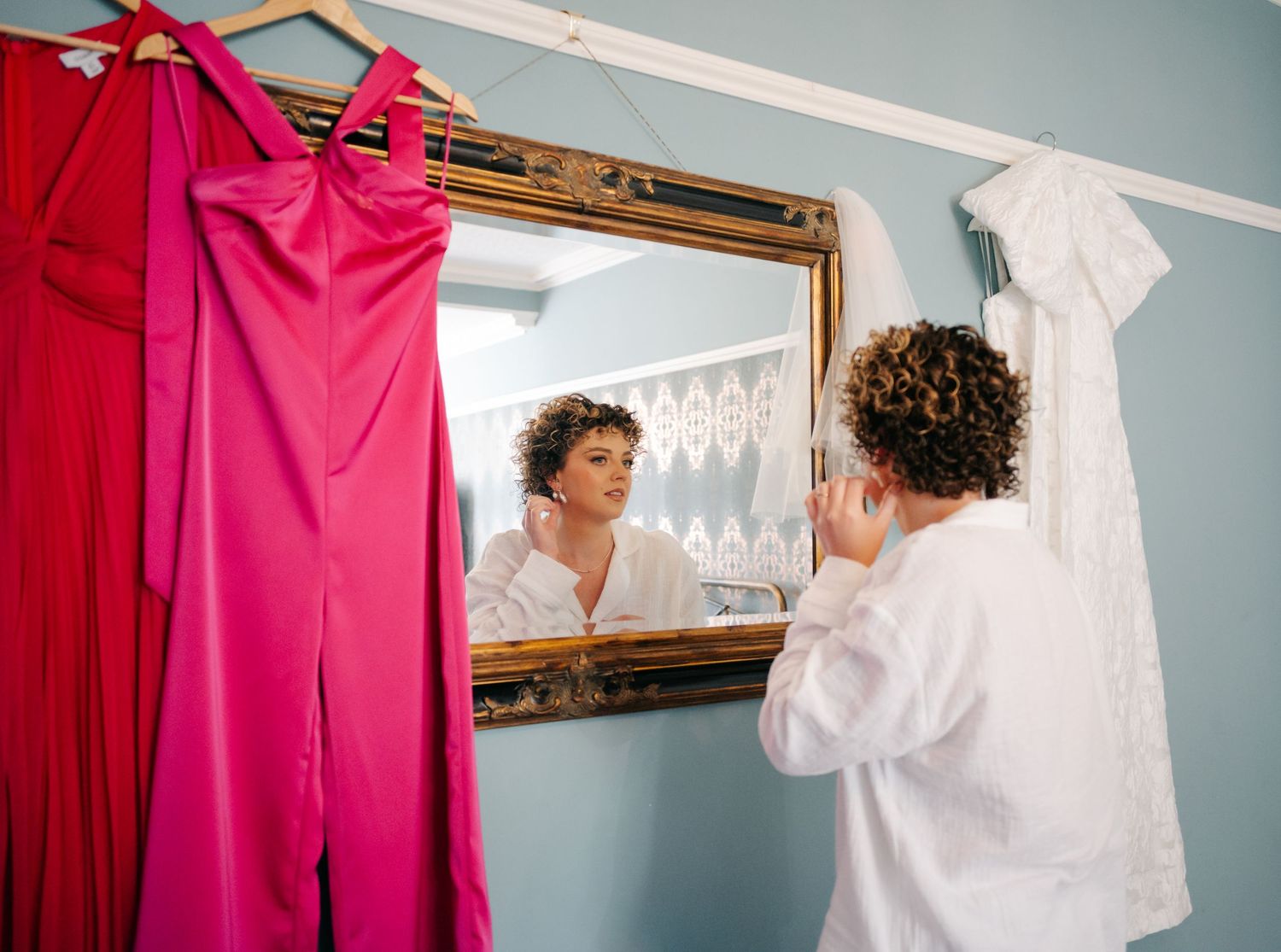 A bright pink dress hangs beside a mirror on a blue wall as someone in white prepares for a special occasion.