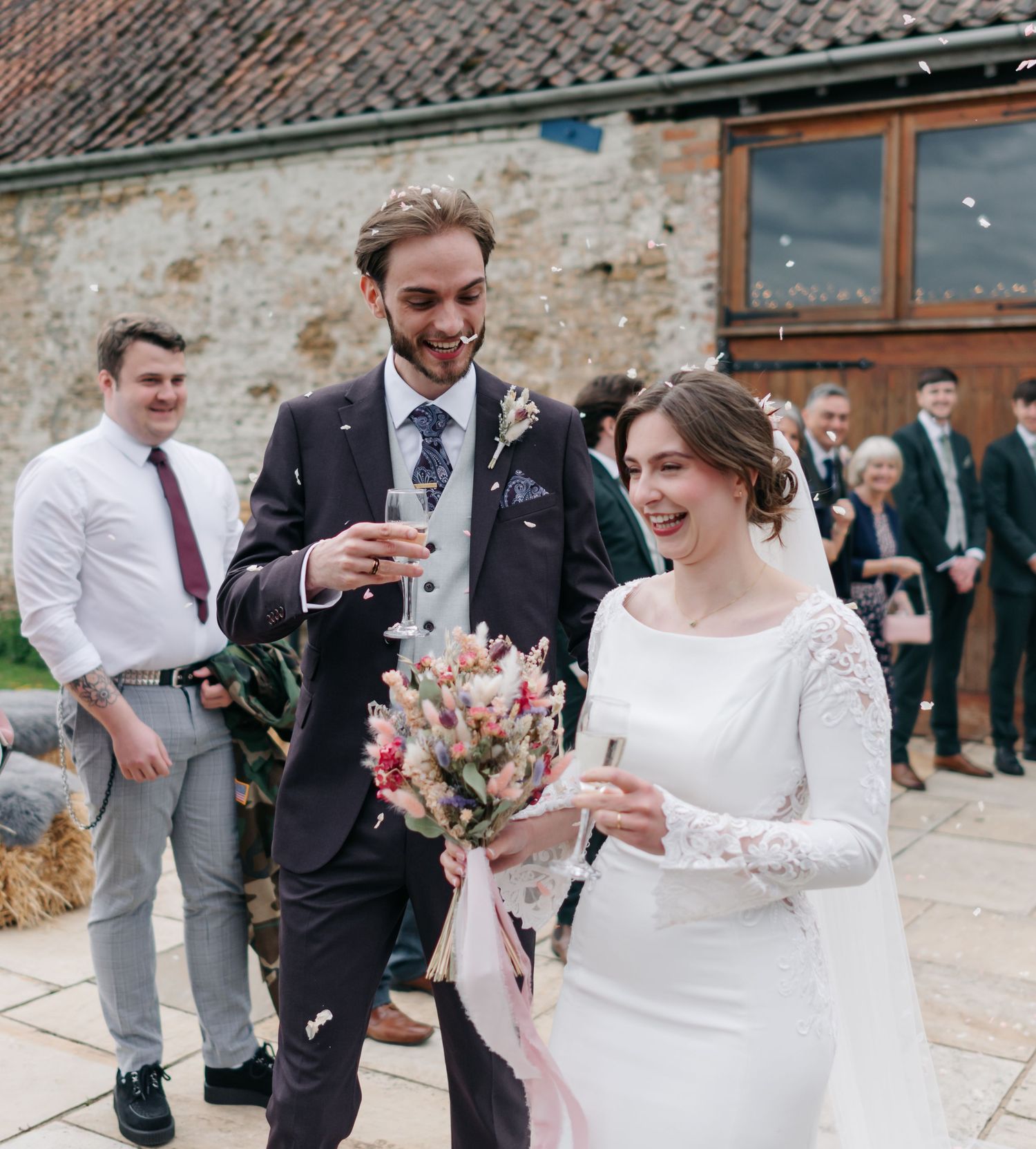 Wedding party members celebrate outside a rustic brick building with joyful expressions.