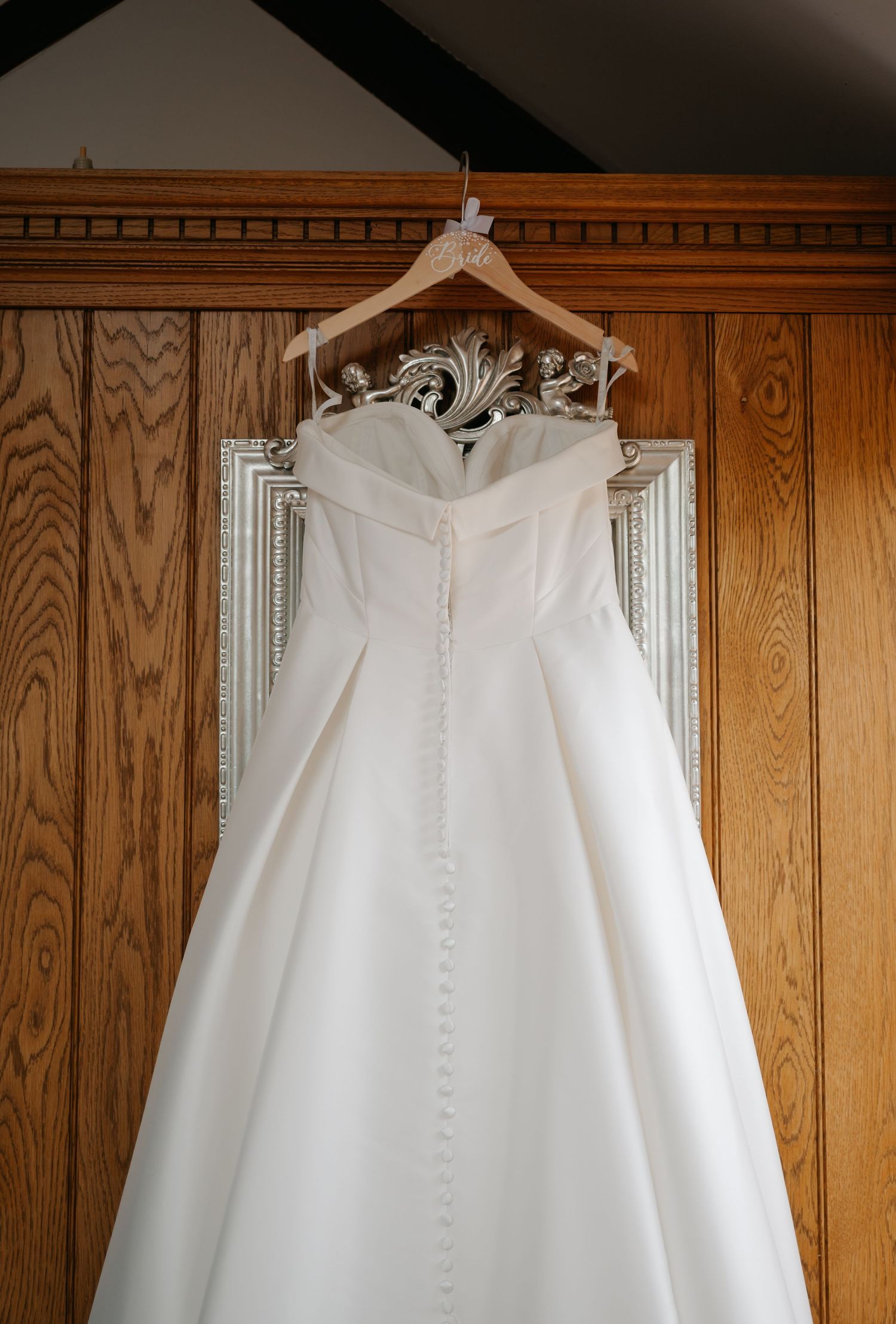 An elegant white wedding dress hanging on a wooden hanger against a rustic oak-paneled wall in natural light.