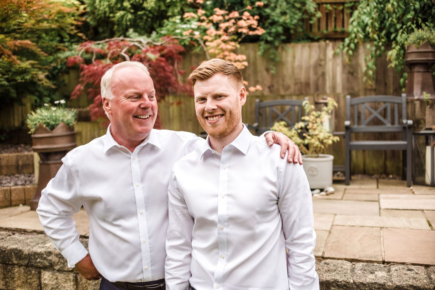Two men in white dress shirts share a joyful moment in a garden setting.