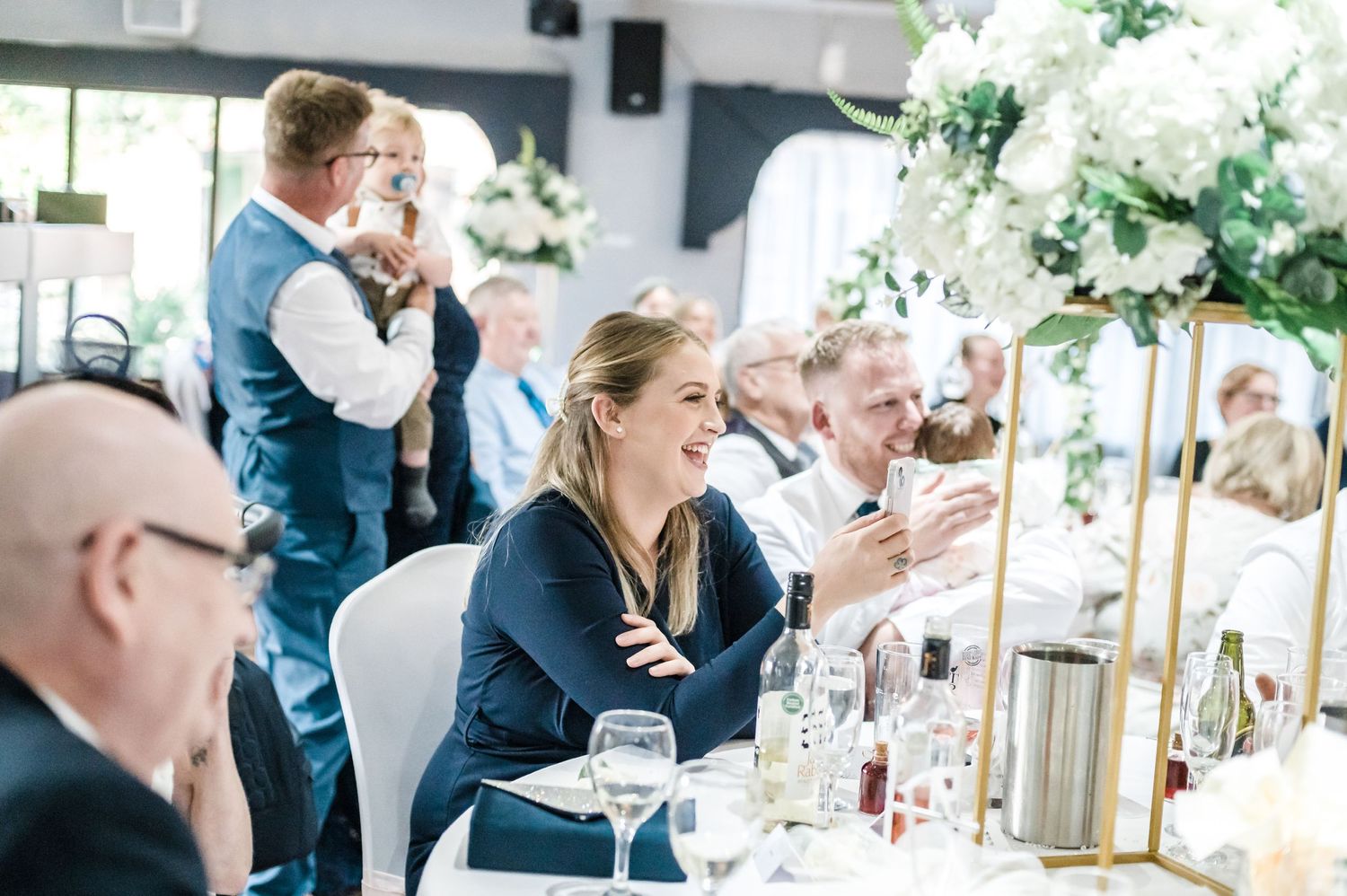Wedding reception scene with guests seated at decorated tables with white floral centerpieces.
