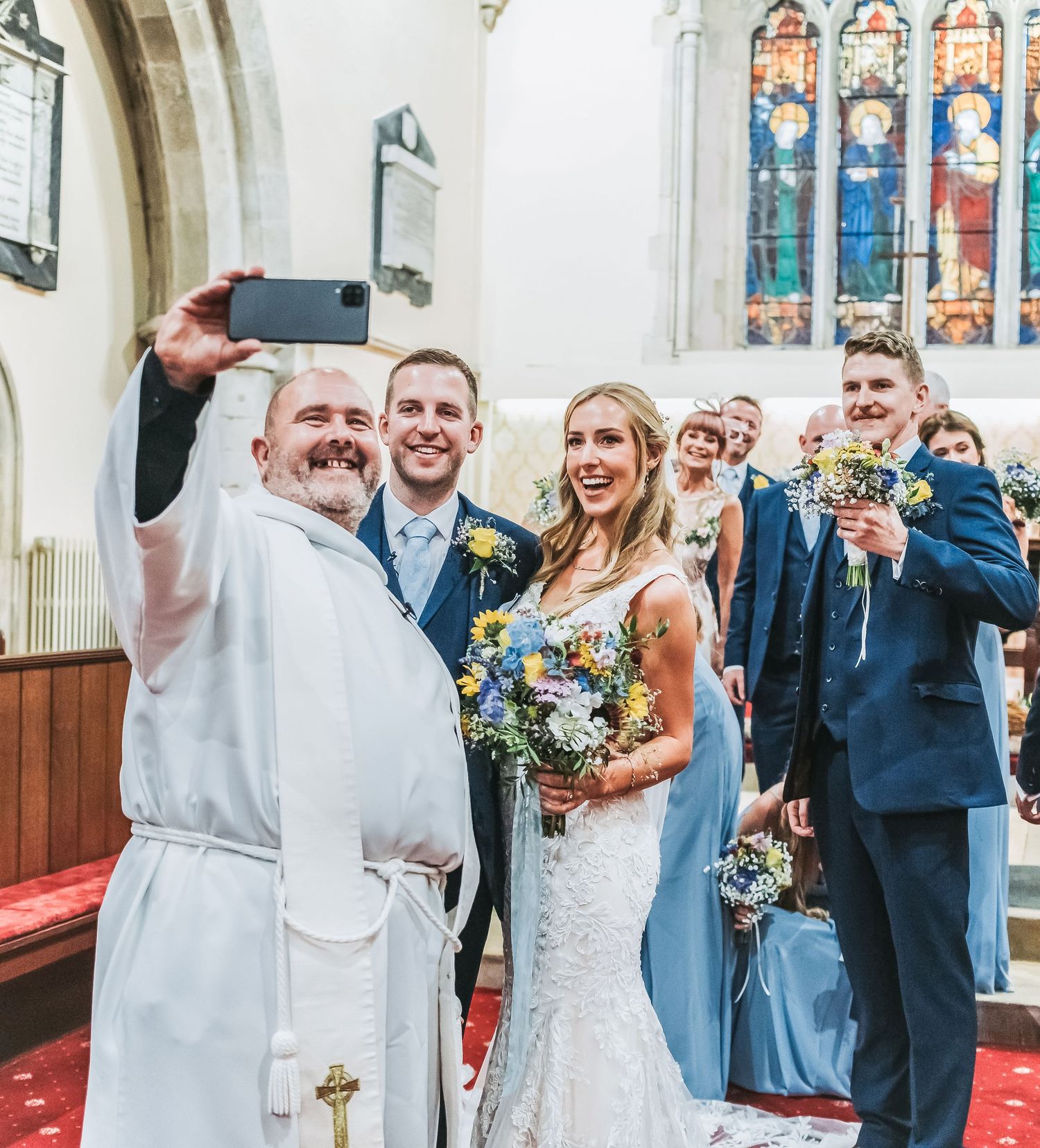Church wedding selfie moment with clergy and wedding party in traditional setting.
