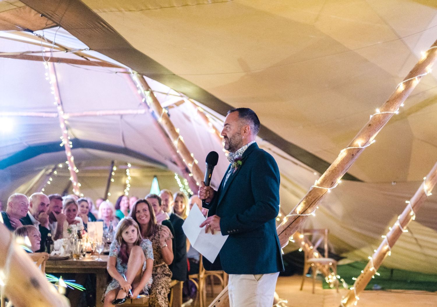 A speaker addressing guests at a tented wedding reception with string lights and decorative ambiance.