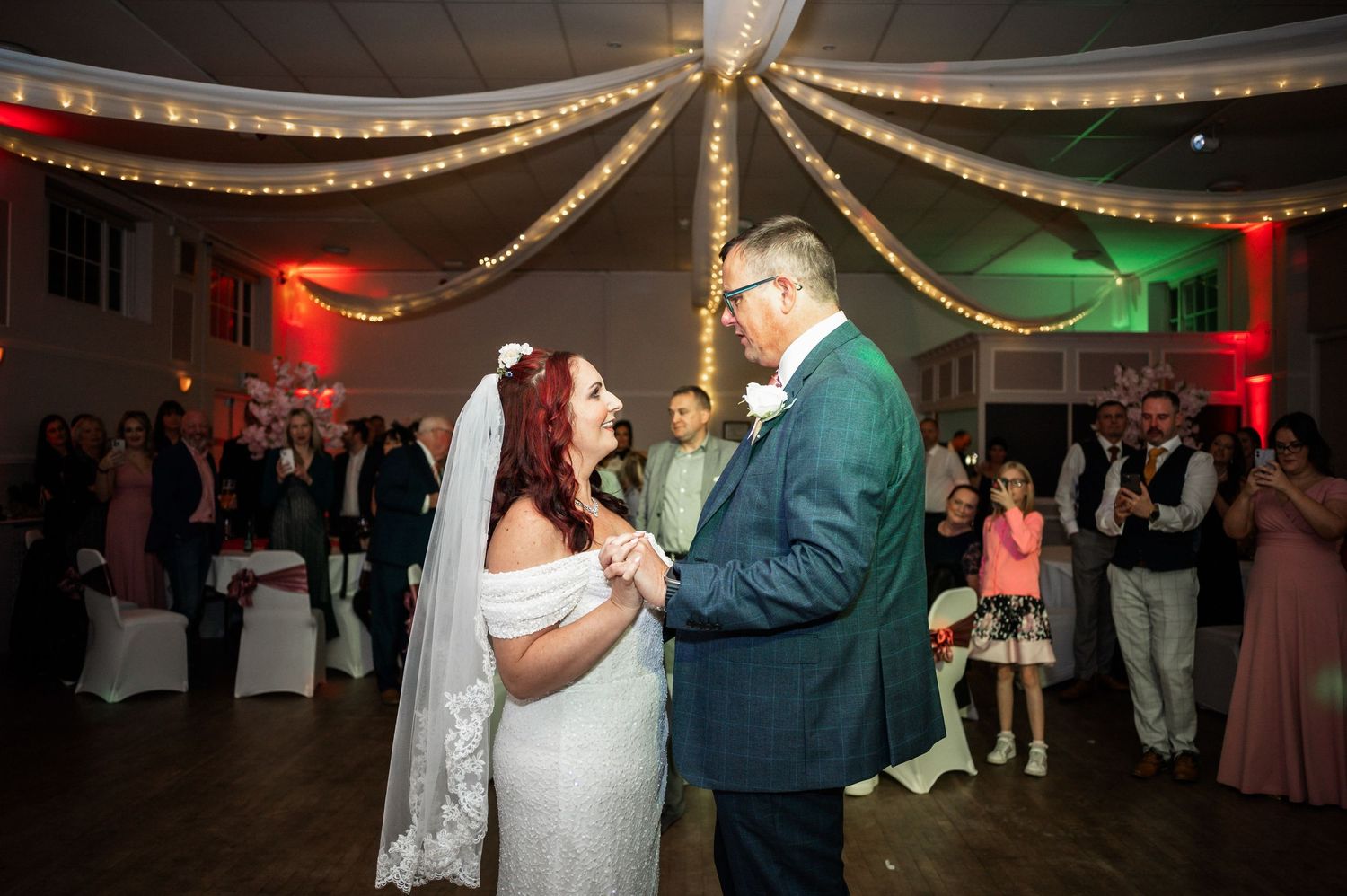 A romantic first dance at a wedding reception under twinkling string lights with red and green accent lighting.