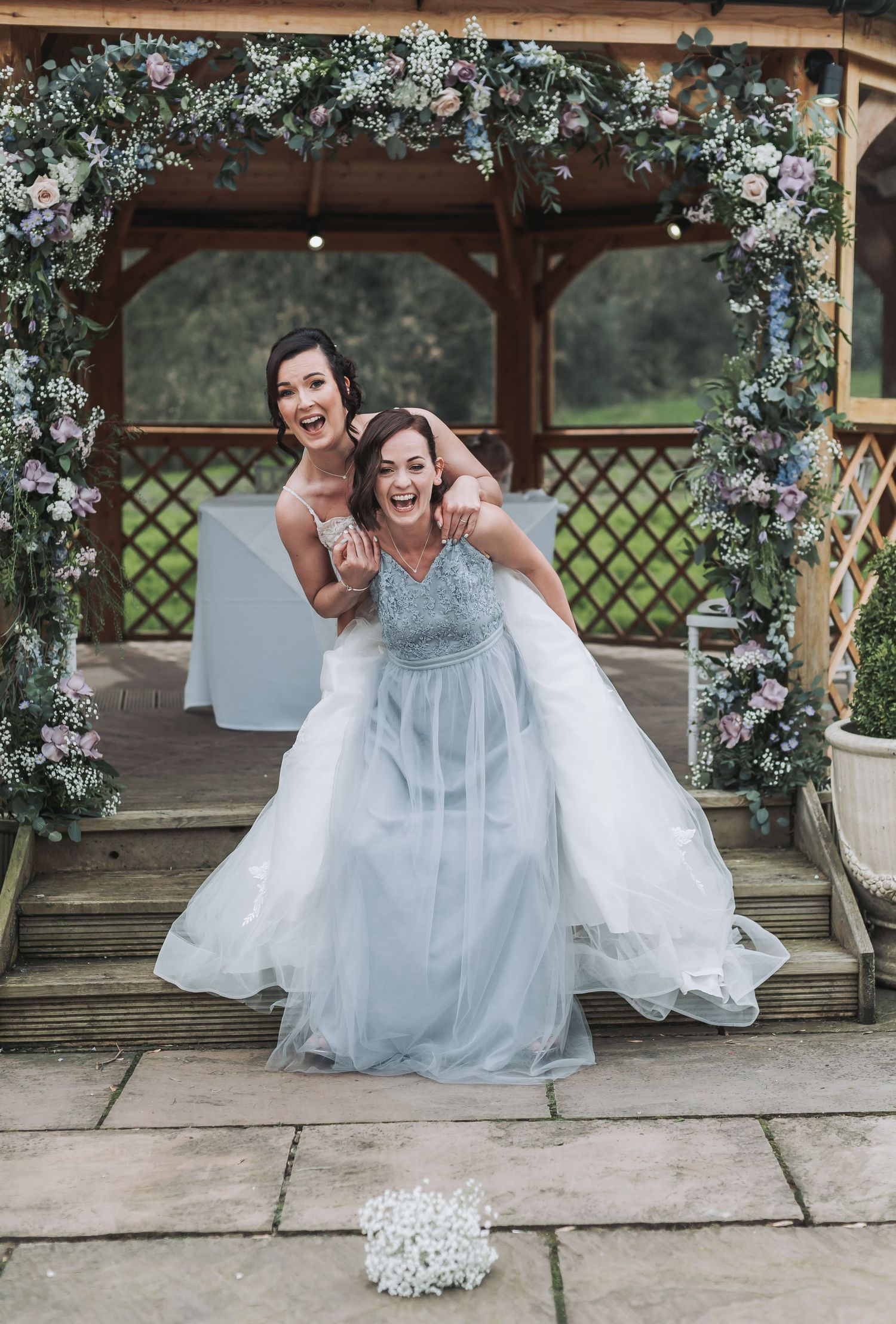 Two bridesmaids in light blue dresses pose playfully under a floral arch at an outdoor wedding gazebo.