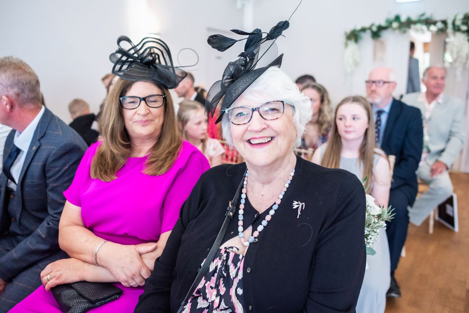Wedding guests seated indoors wearing festive hats and bright attire at a celebration.
