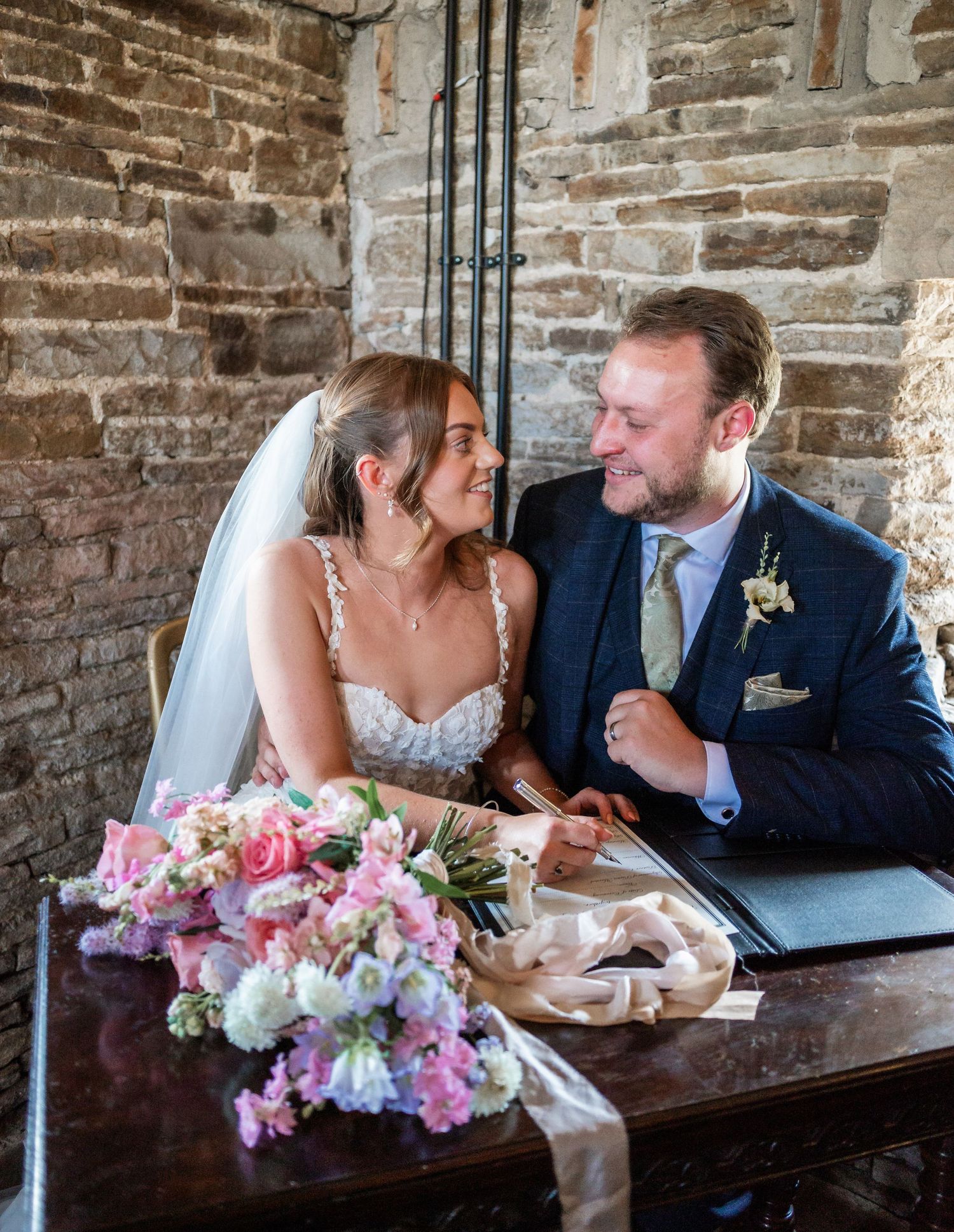 Romantic wedding couple shares intimate moment at antique table with pink and white flower bouquet.