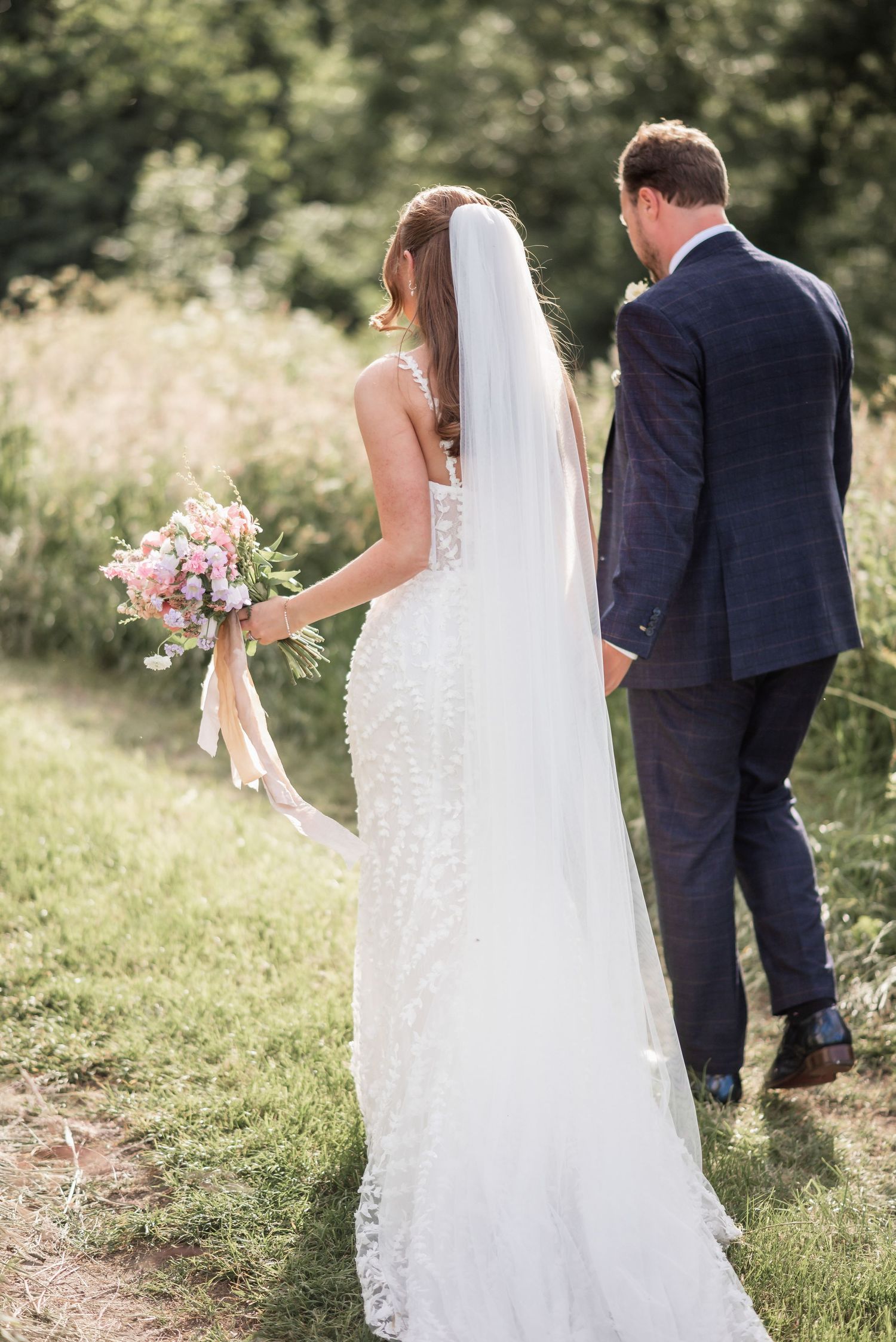 A couple walks through a sunlit meadow during their wedding photoshoot.
