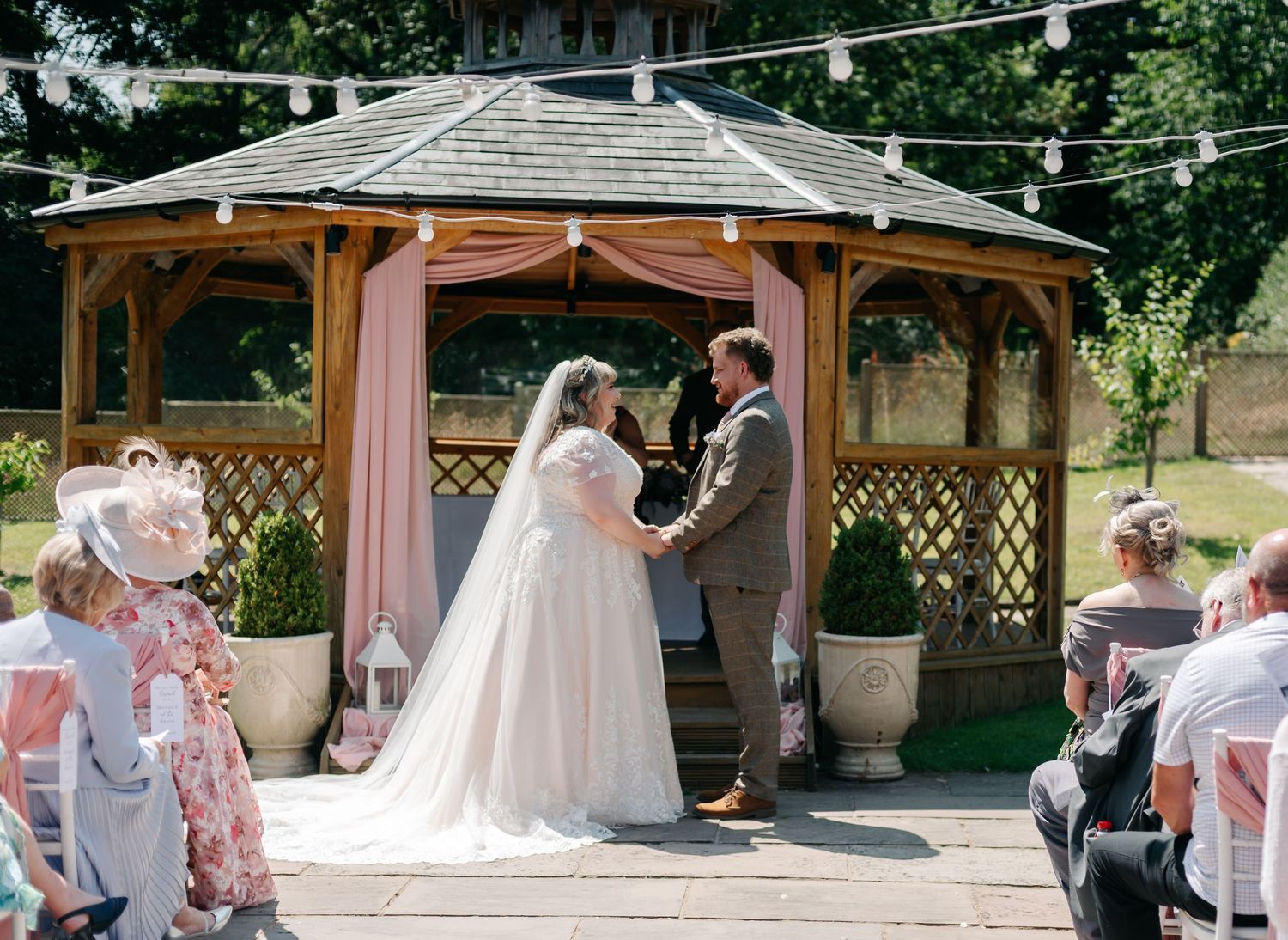 Bride and groom exchange vows at an outdoor gazebo wedding ceremony decorated with pink draping on a sunny day.
