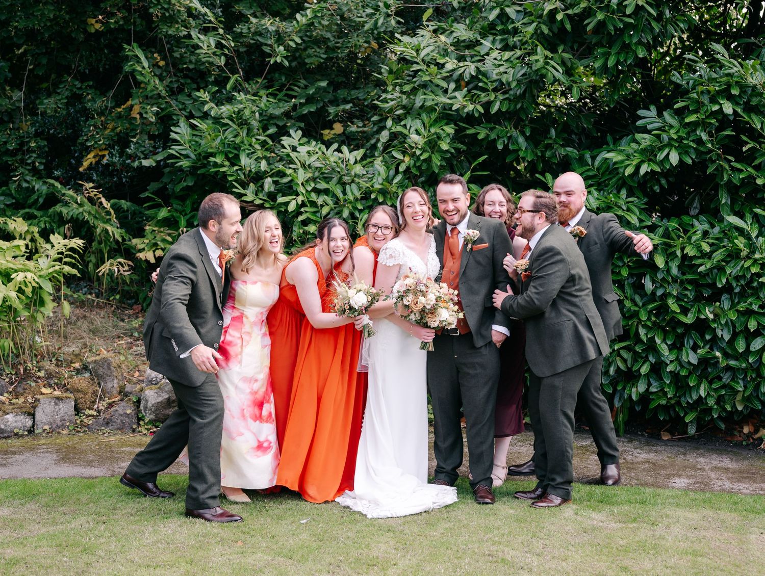 Bride in a white gown and bridesmaids in coral and orange dresses laugh with groomsmen in suits during a joyful outdoor wedding group photo.